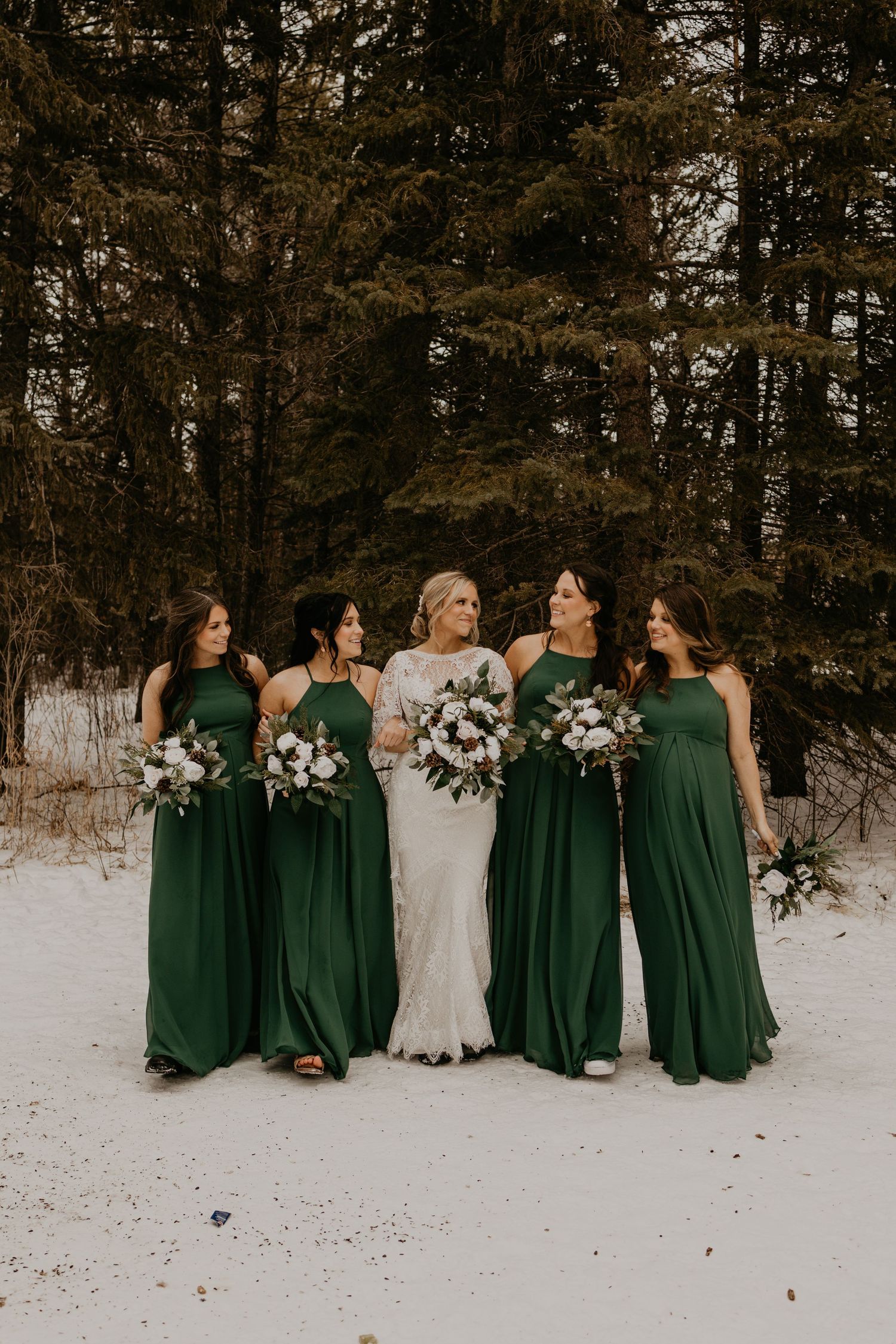 Wedding party in emerald and white gowns poses together on snowy ground with pine trees in background.