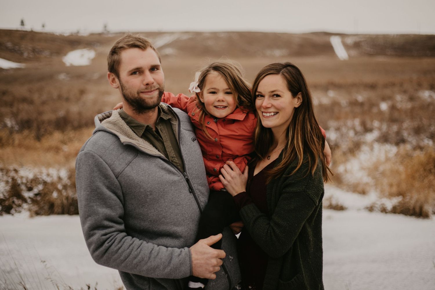 A warm family portrait outdoors with snow patches and golden grass in the background.