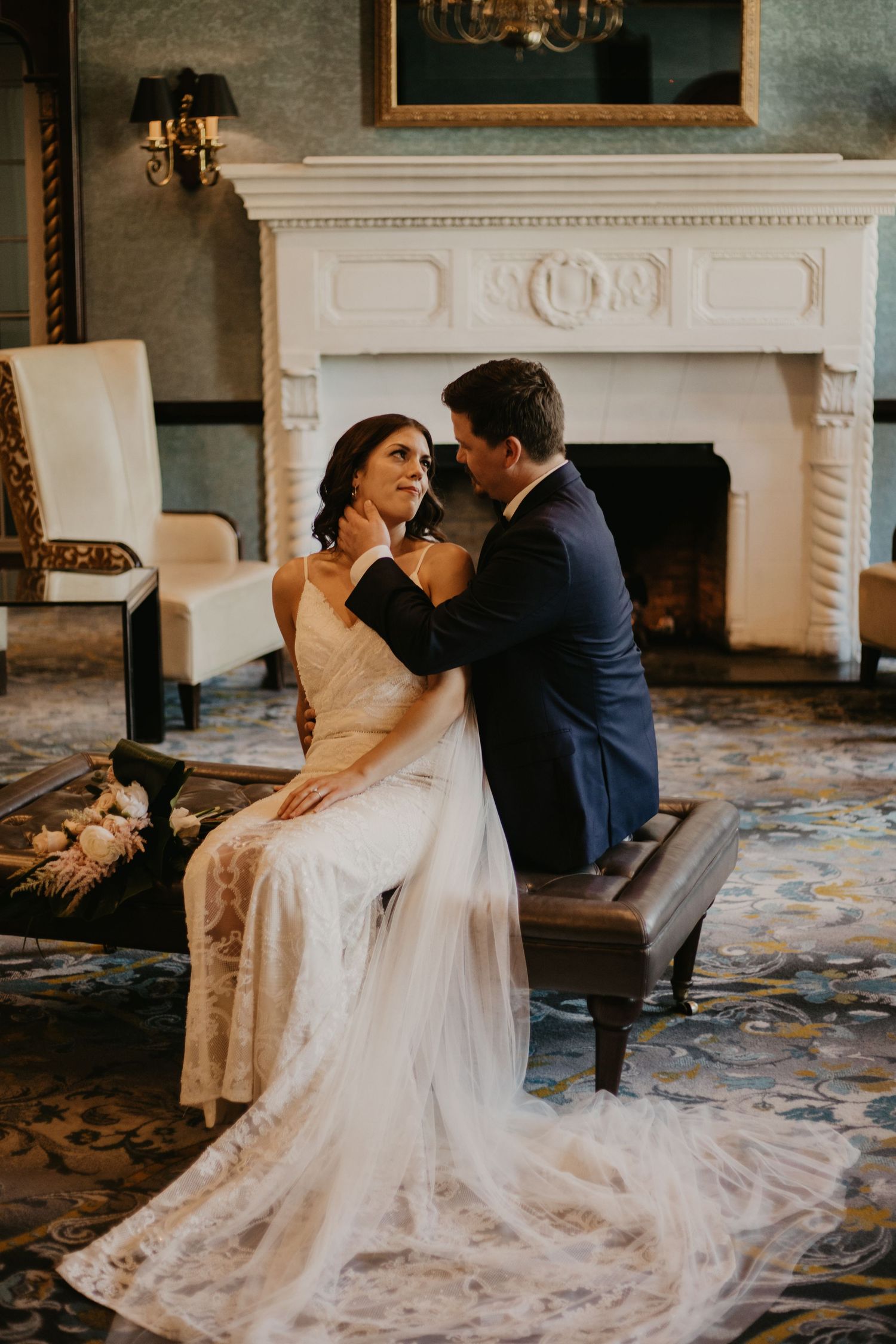 A romantic wedding moment captured in front of an ornate fireplace with a bride's flowing lace gown trailing behind.