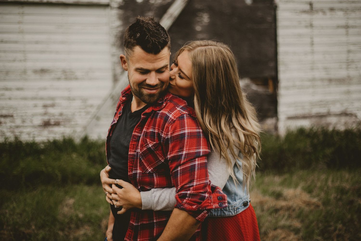 A couple sharing an intimate moment by a weathered barn wall in casual fall attire.