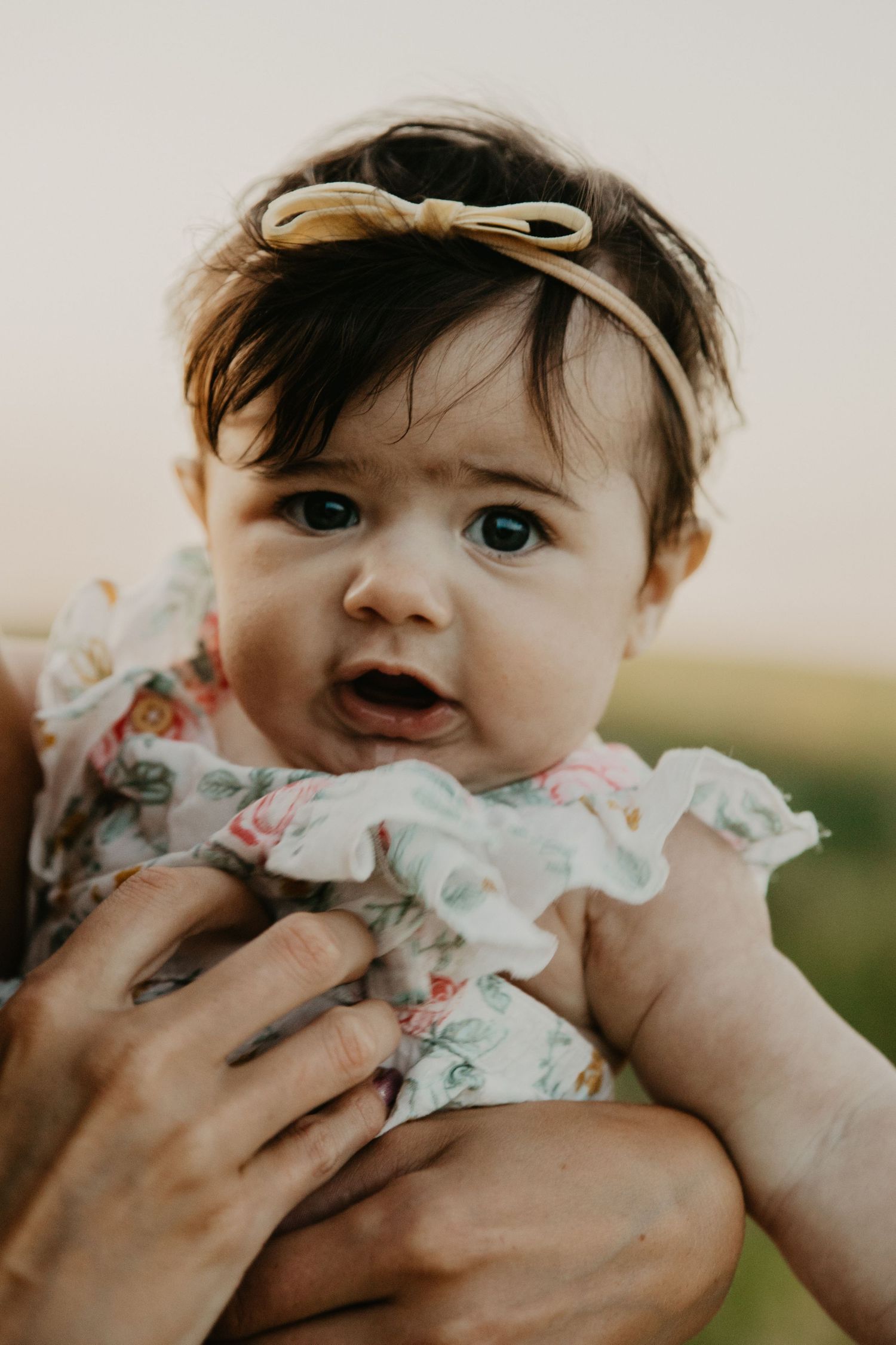 Baby wearing floral dress with bow headband being held by gentle hands against neutral background.