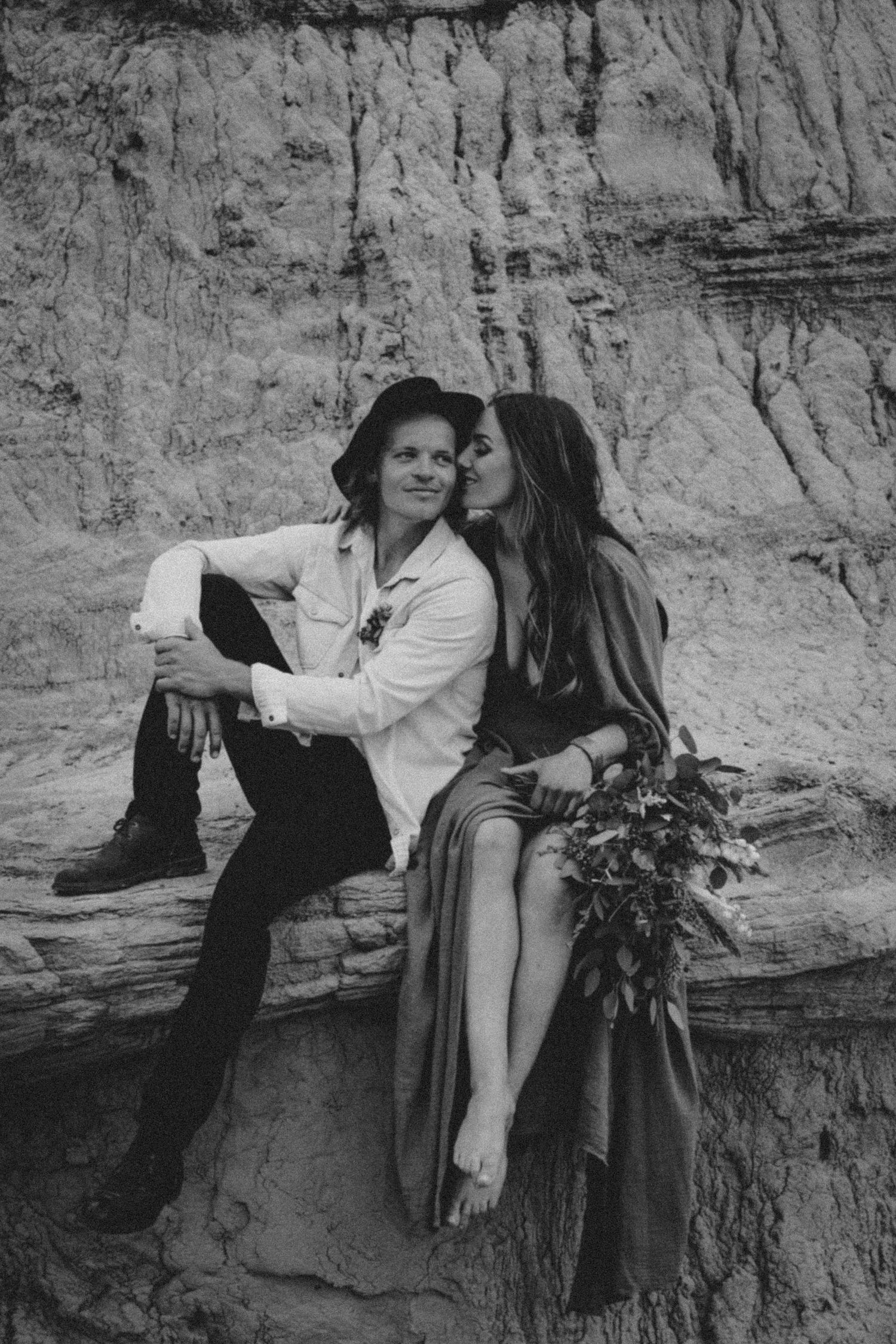 A black and white portrait of a couple sitting on rocky terrain with wildflower bouquet.