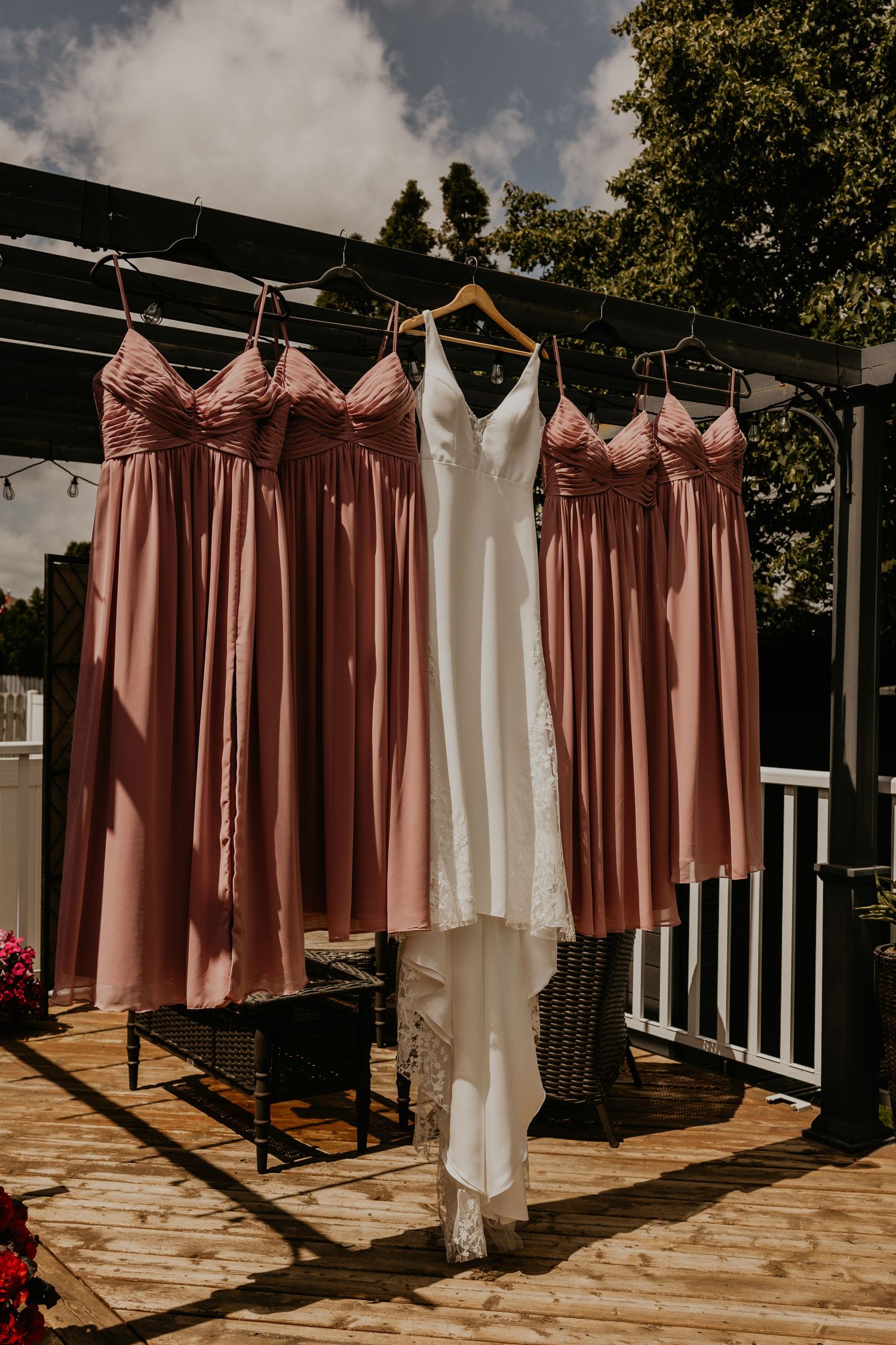 A white wedding dress and pink bridesmaid dresses hanging on a wooden pergola outdoors.