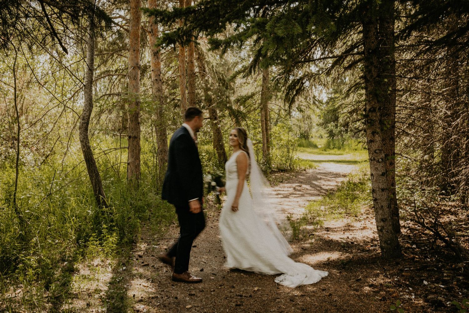 Wedding couple walking on forest path surrounded by evergreen trees.
