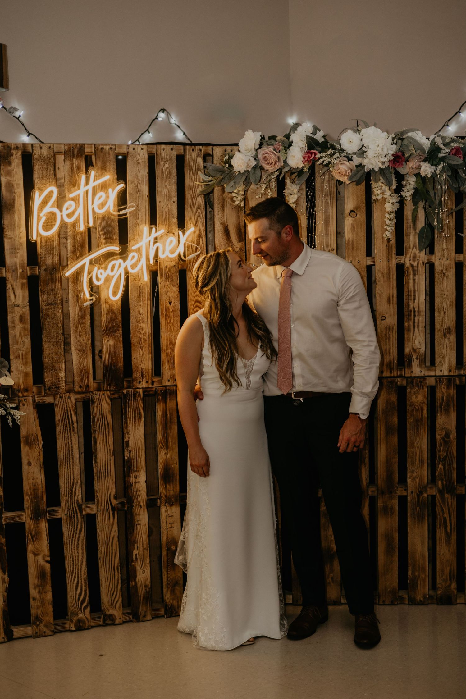 A romantic moment between newlyweds in front of a wooden pallet wall with neon 'Better Together' sign.