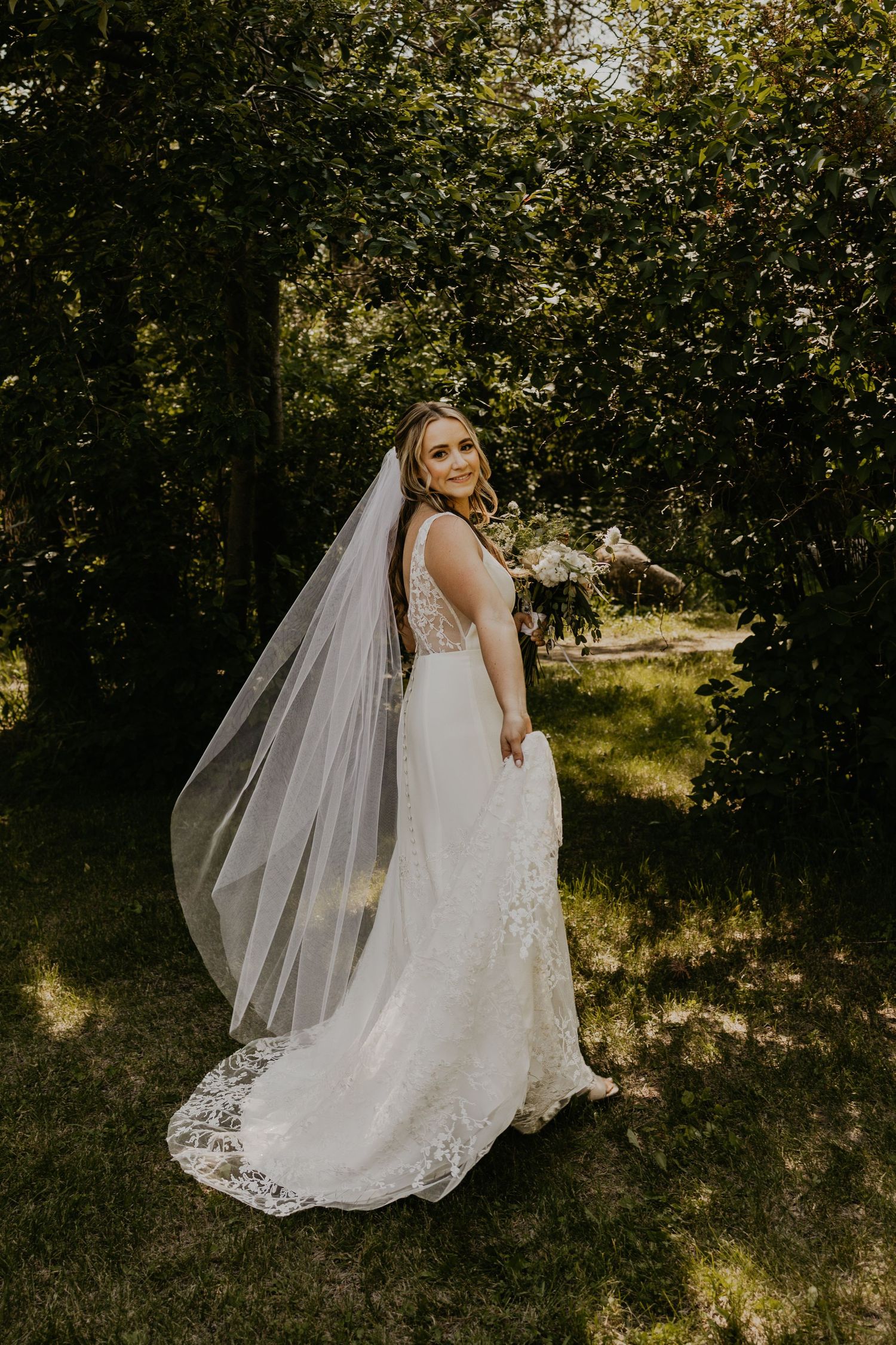 Bride in lace-detailed gown with flowing veil poses against wooded background.