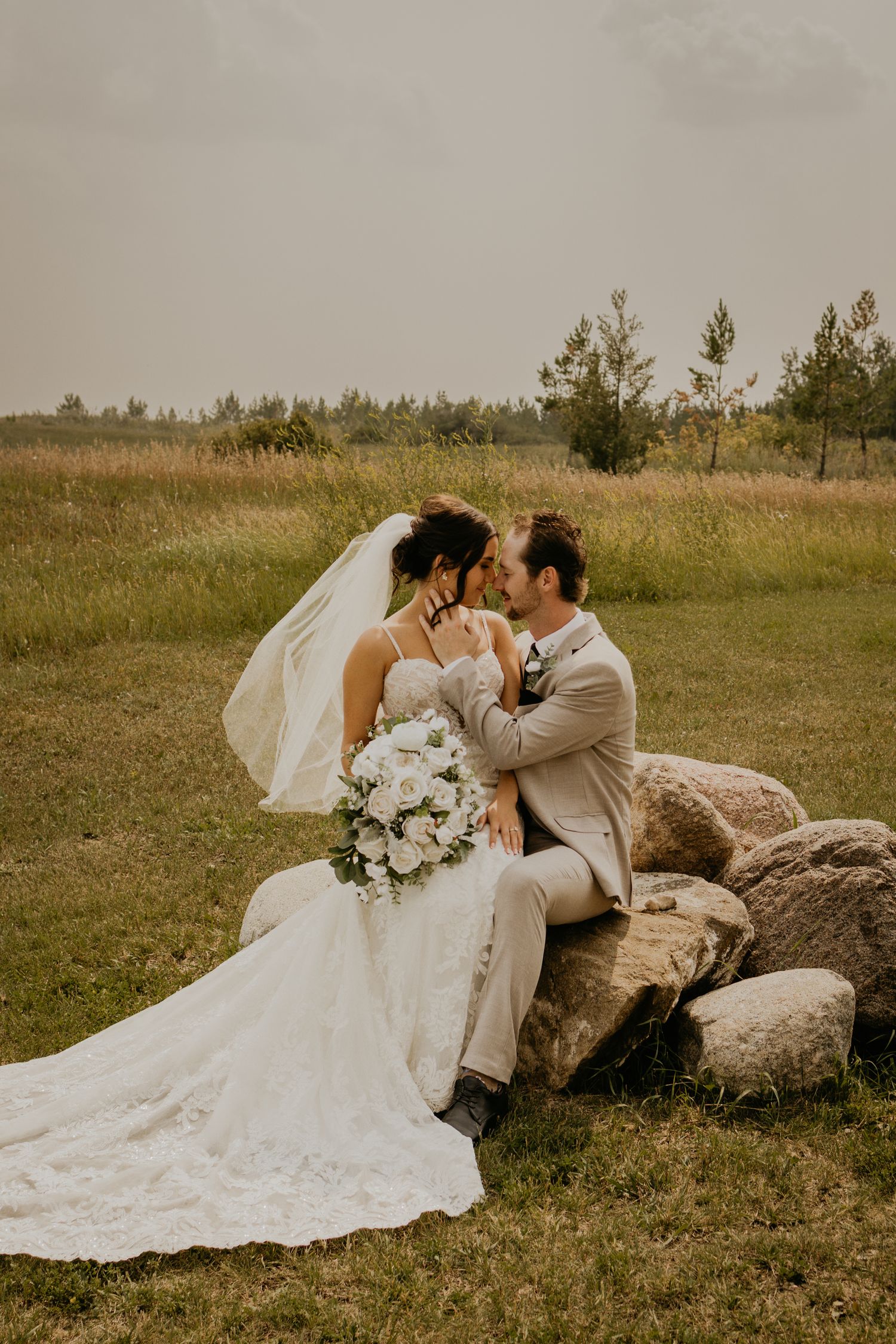 A wedding couple shares an intimate moment while sitting on rocks in a rustic outdoor field setting at sunset.