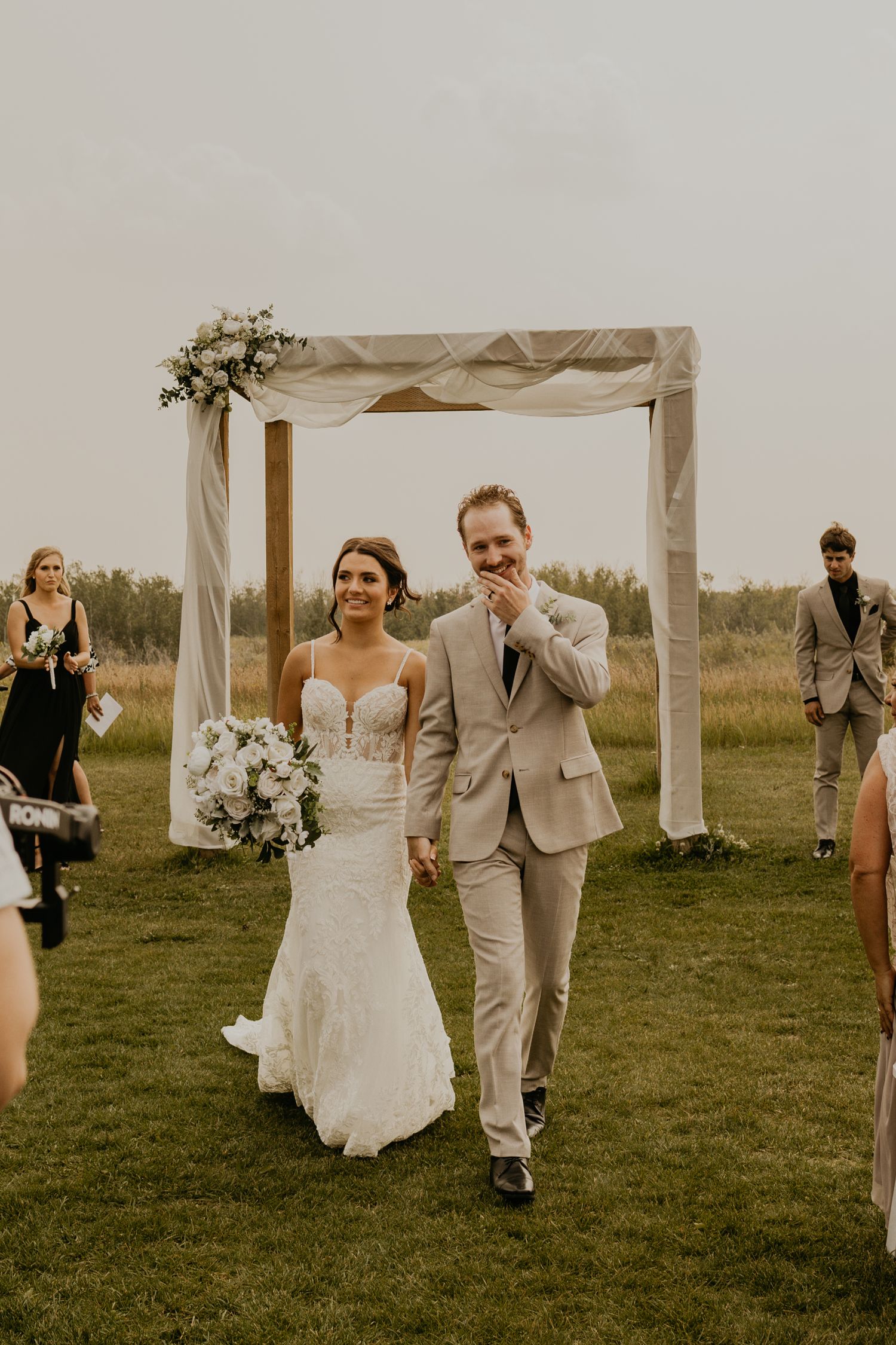 Newlyweds exit their ceremony under wooden arch decorated with white fabric and flowers.