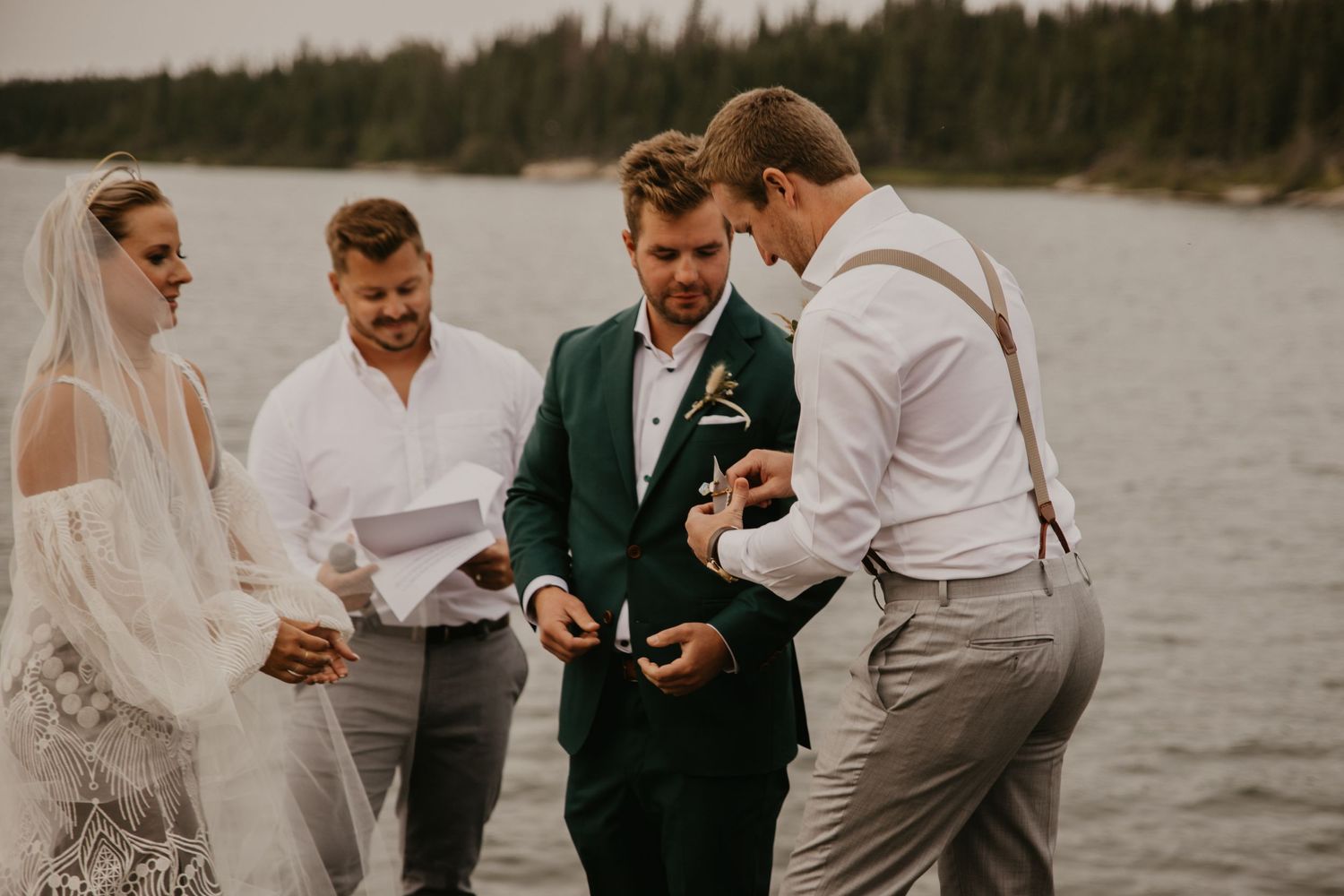 Small wedding ceremony taking place by lakeside with forest in background.