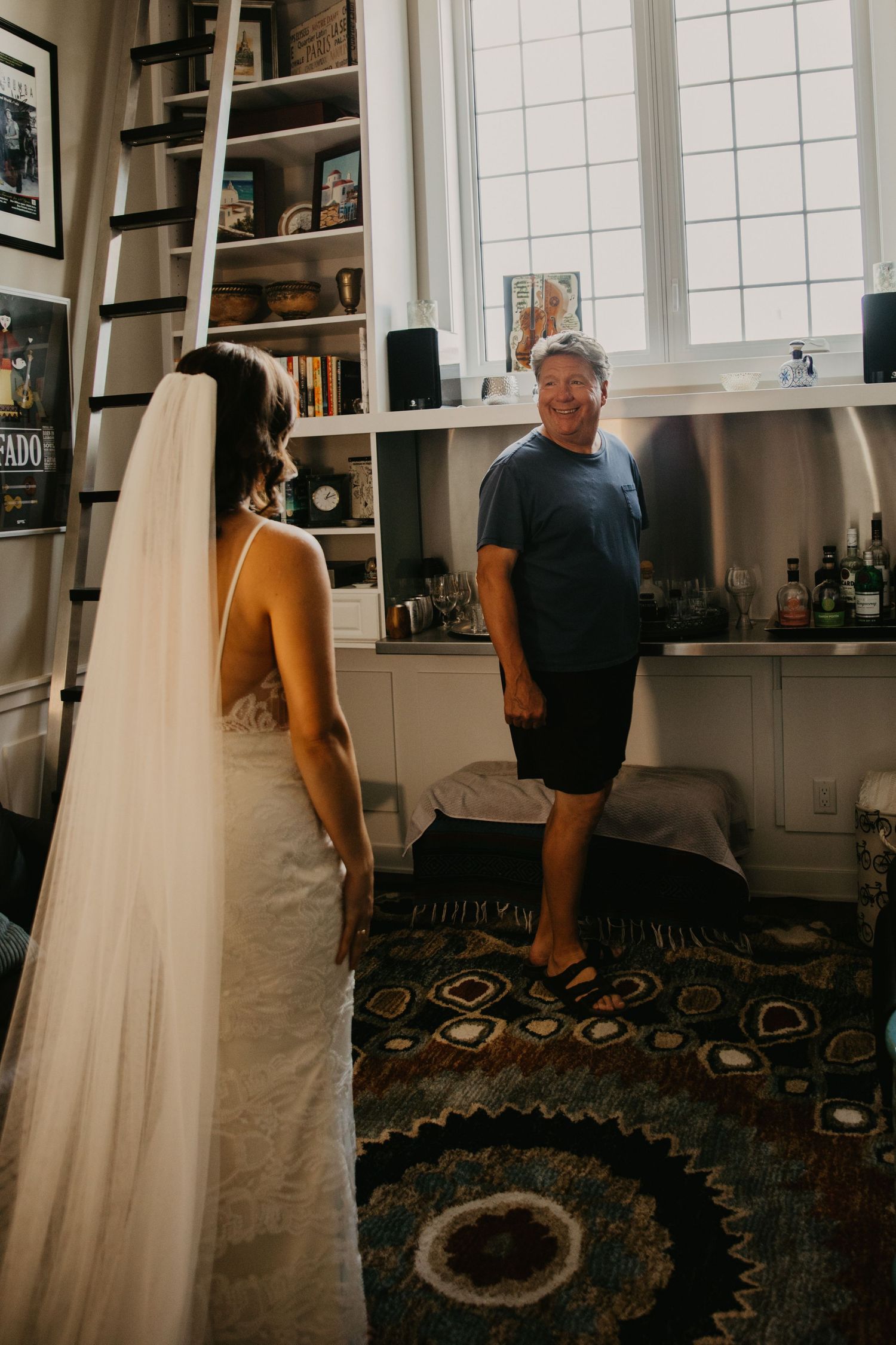 A bride in a white wedding dress shows her gown while standing on a patterned rug in an intimate home setting.