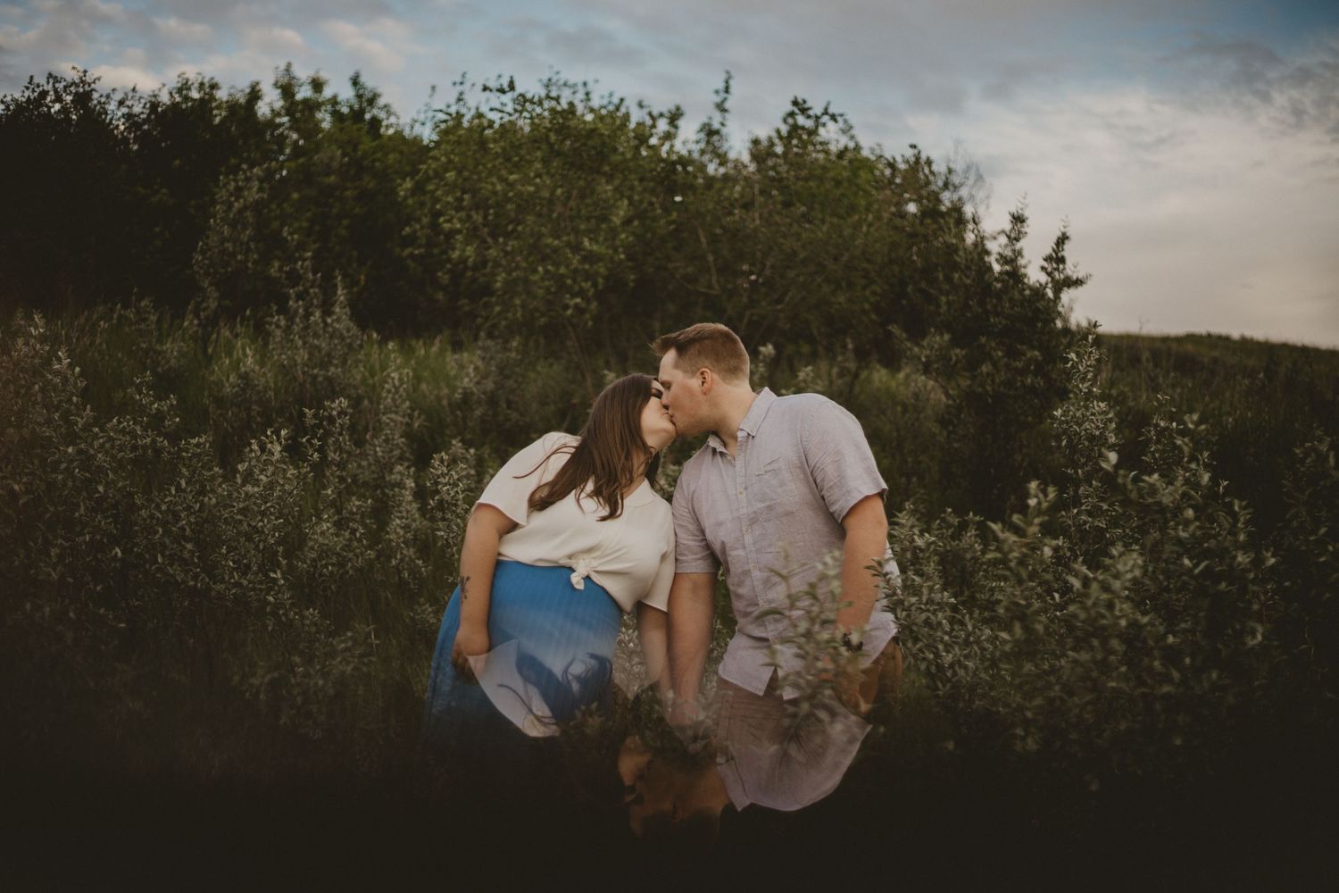 A couple shares an intimate moment among green foliage at dusk.