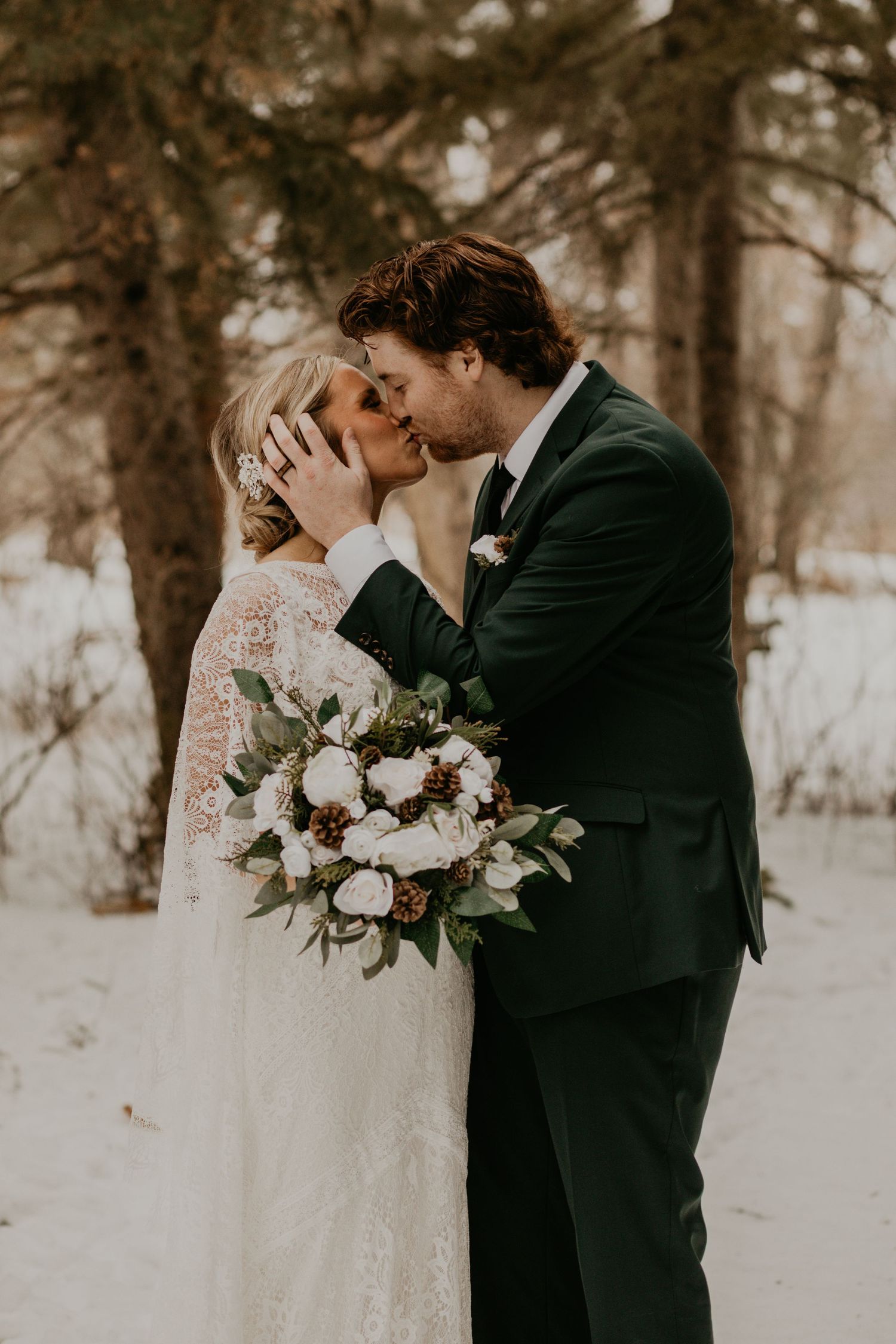 Romantic winter wedding kiss captured in a forest with a beautiful white floral bouquet.