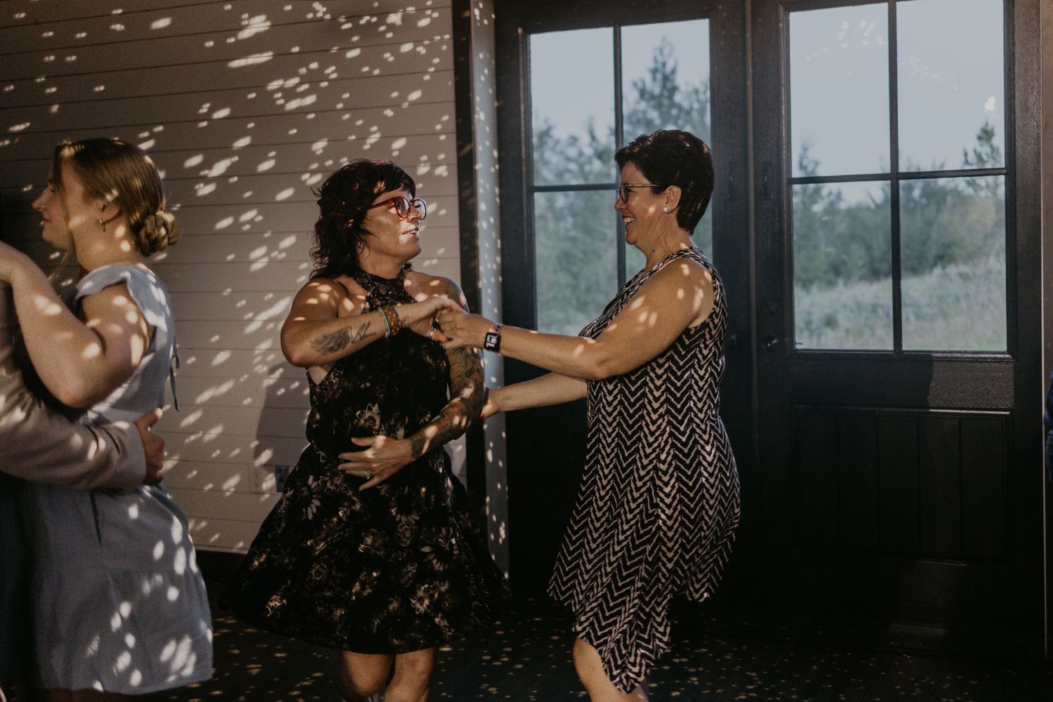 Reception guests dance together under sparkling lights streaming through windows.