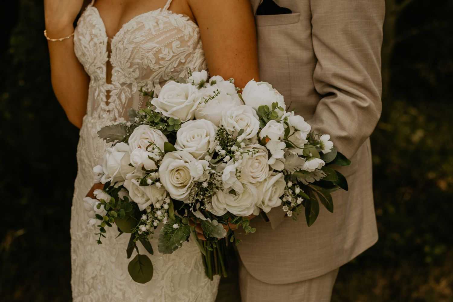 Close-up of elegant white rose wedding bouquet held against lace dress and beige suit.