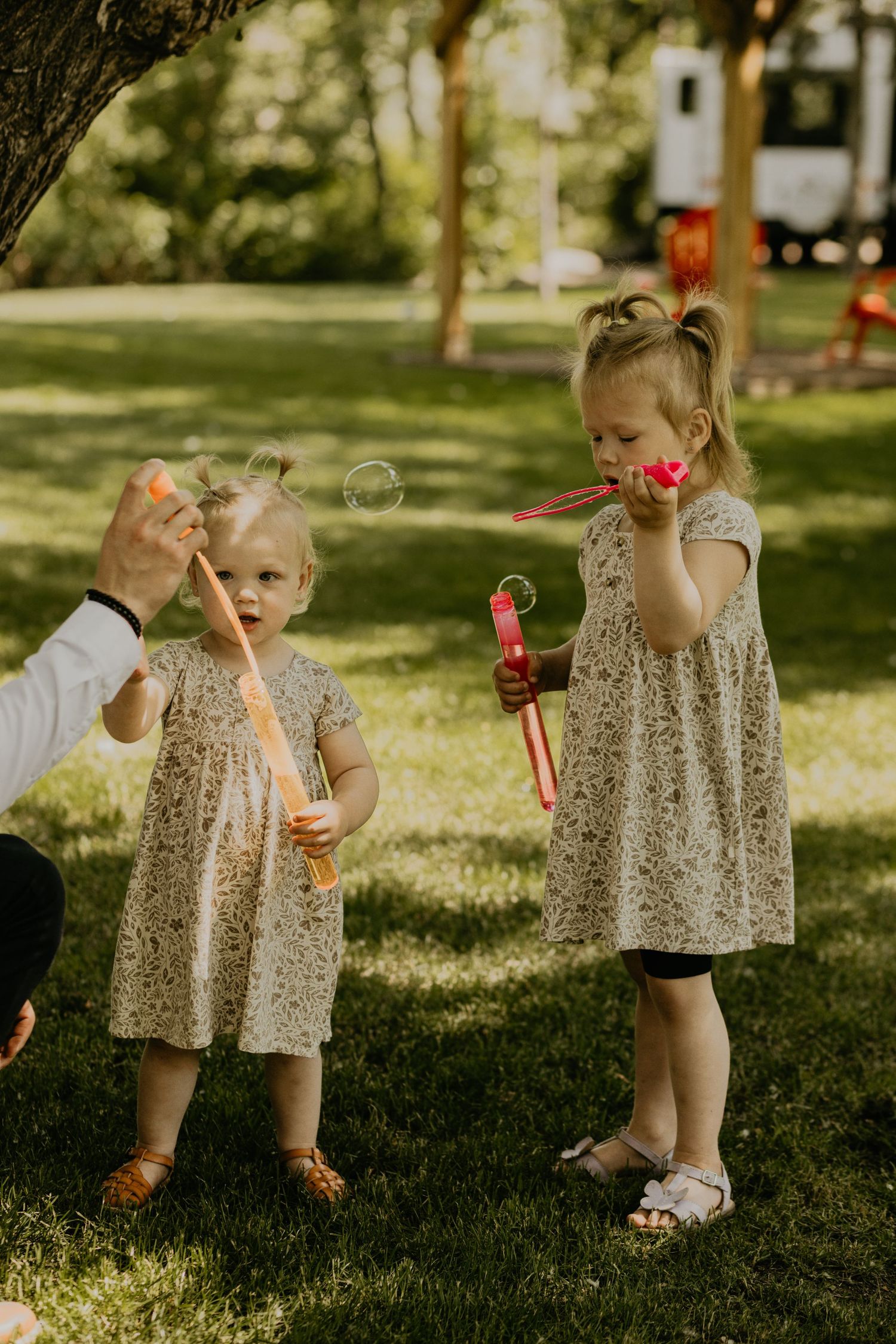 Two young girls in floral dresses play with bubbles on sunny lawn.
