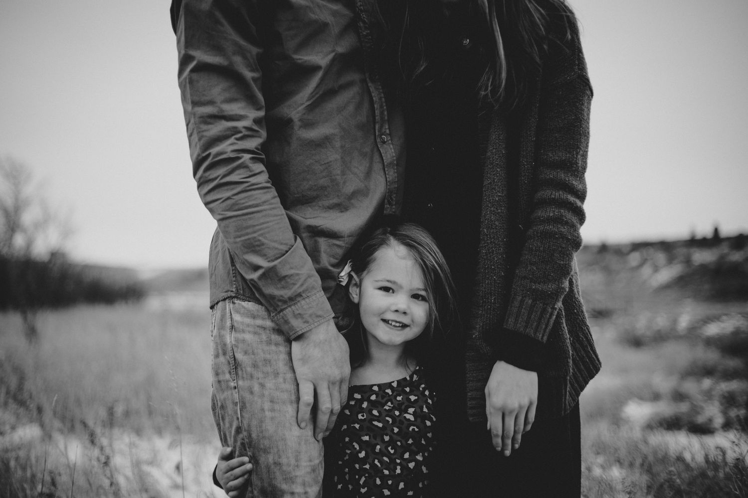 A candid black and white photograph showing a child in a polka dot dress standing between two adults outdoors.