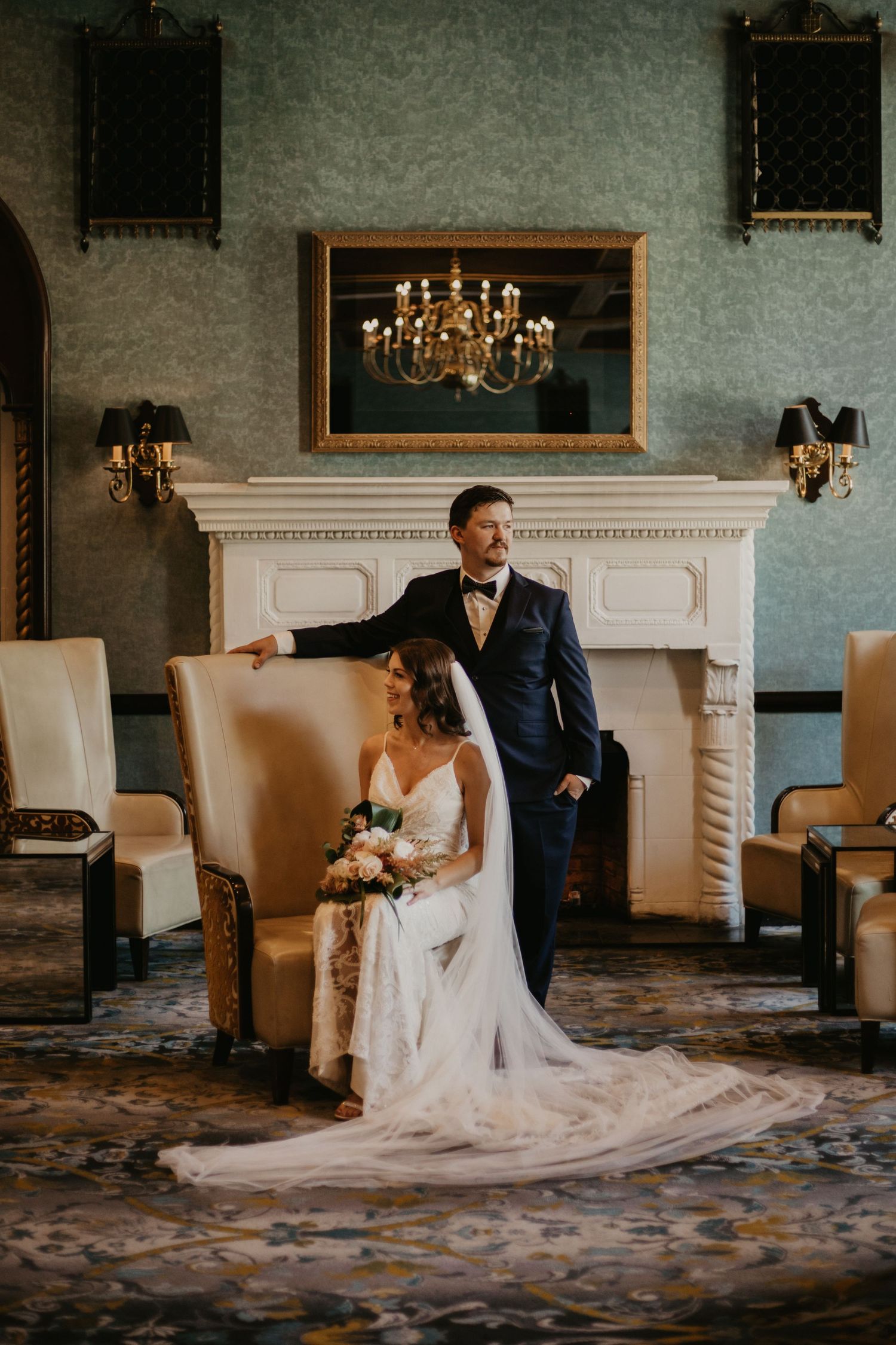 Elegant wedding portrait in front of an ornate fireplace with chandelier reflection.