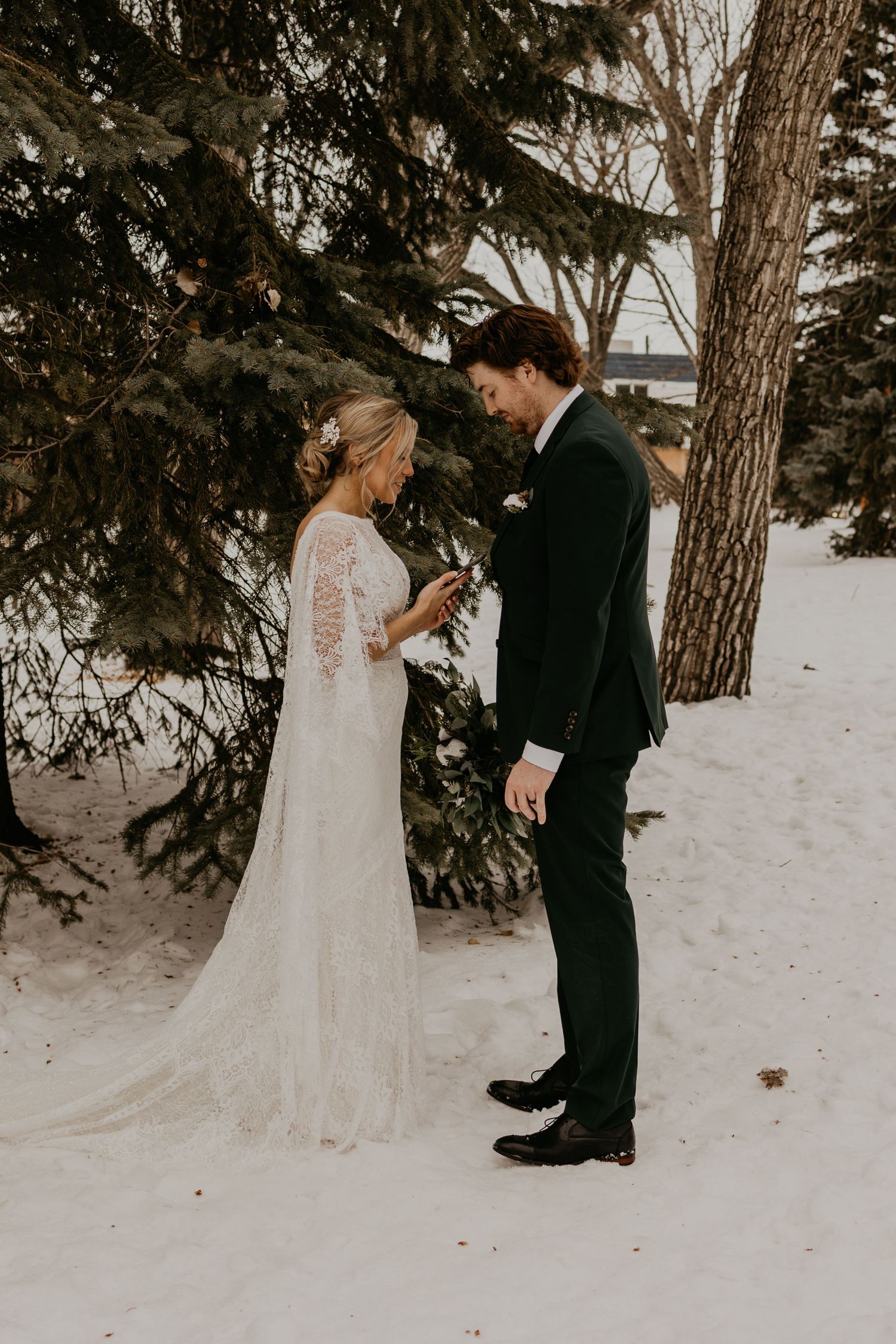 Wedding couple exchanges vows in snowy winter setting beneath evergreen trees.