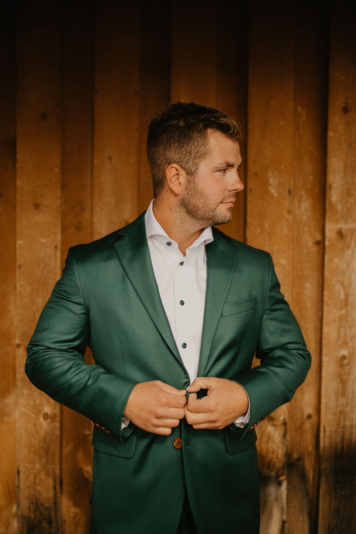 A groom in an emerald green suit jacket adjusts his buttons while standing against a rustic wooden wall.