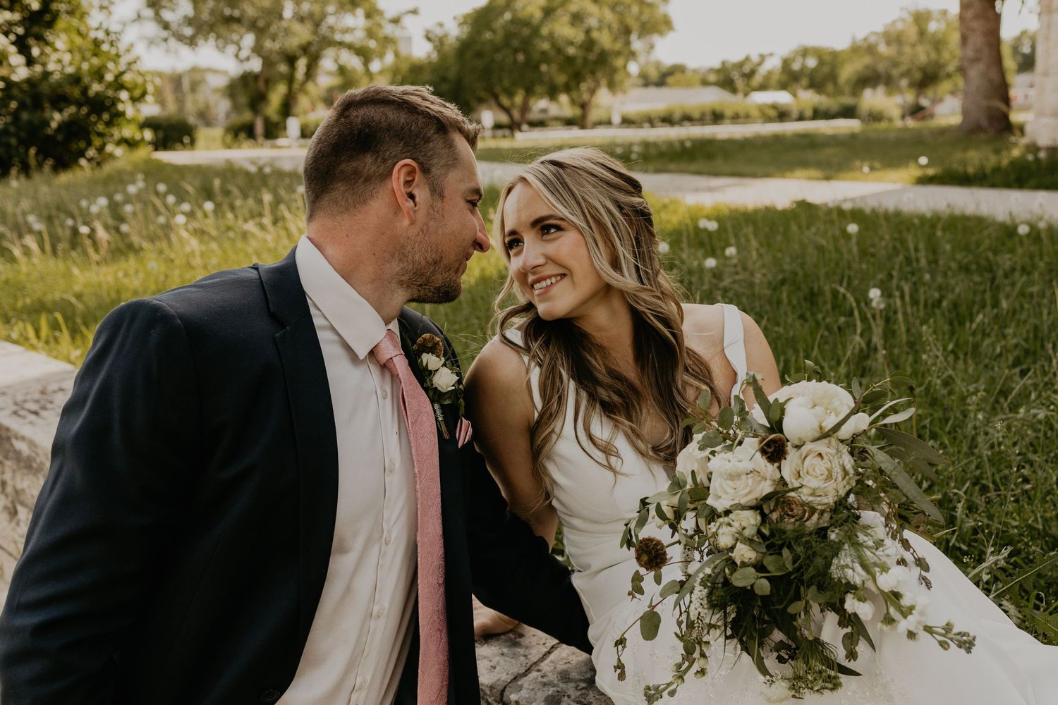 Couple shares romantic moment in sunlit garden setting with white floral bouquet.