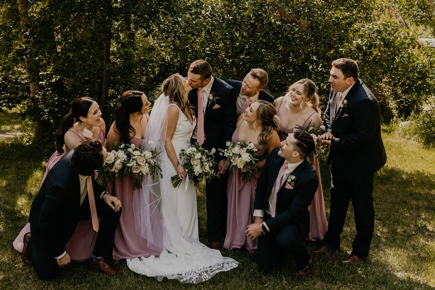 Wedding party shares candid moment while laughing together on sunny lawn.