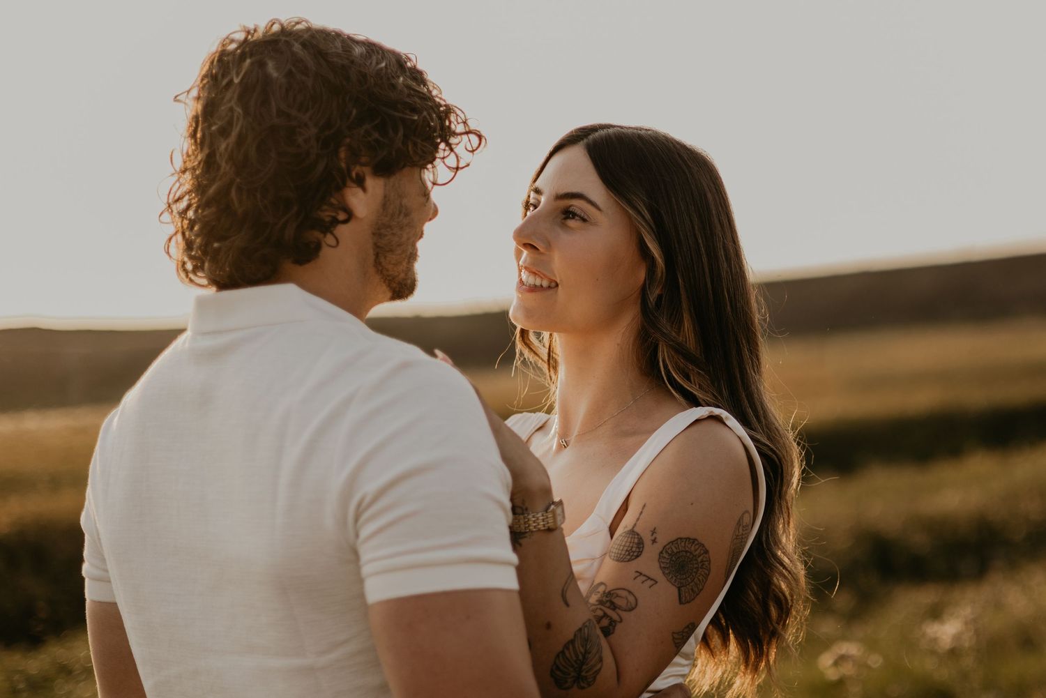 Romantic golden hour photoshoot featuring couple in white outfits against scenic meadow landscape.