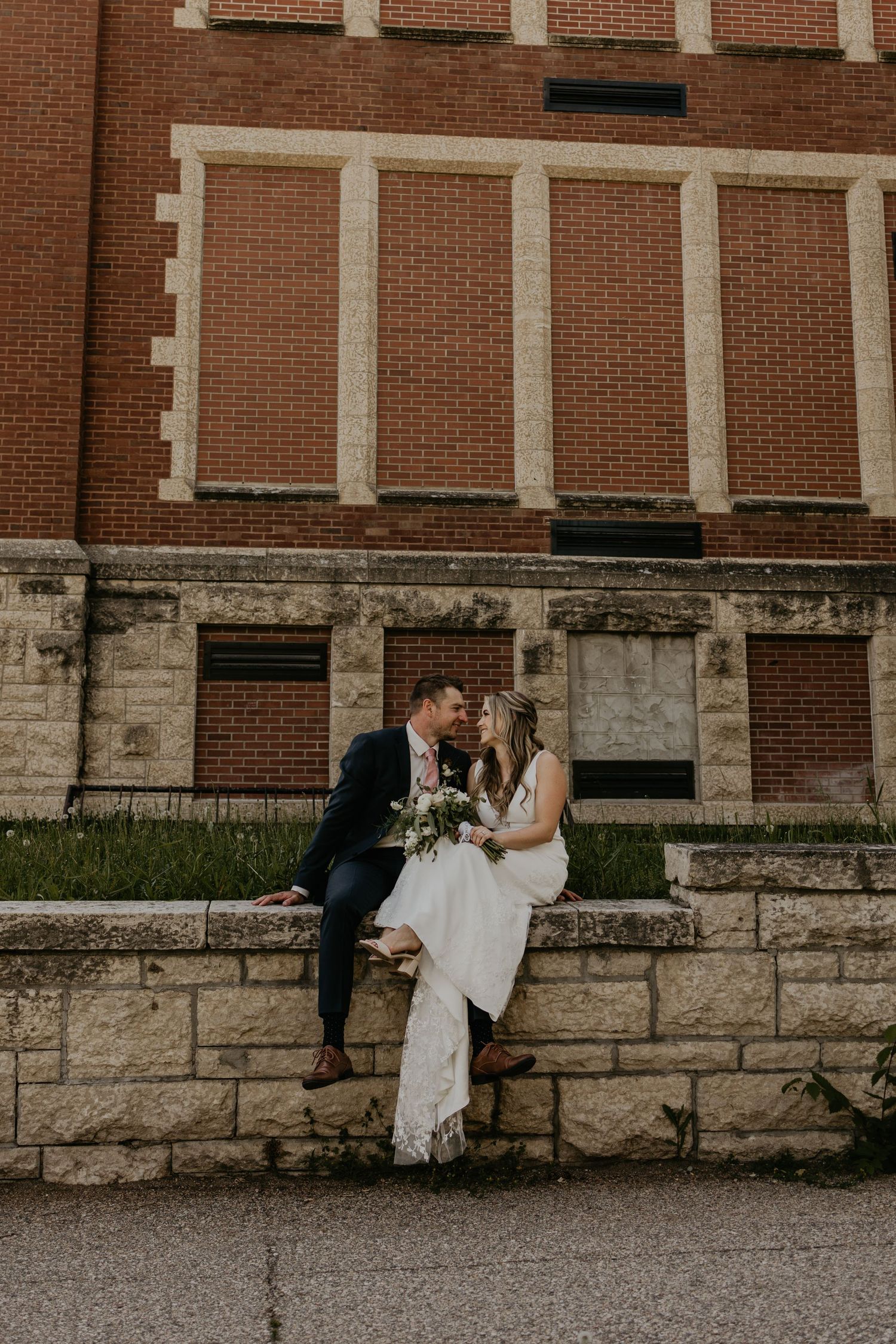 A romantic wedding photo of a couple sitting on stone steps against a historic brick building.