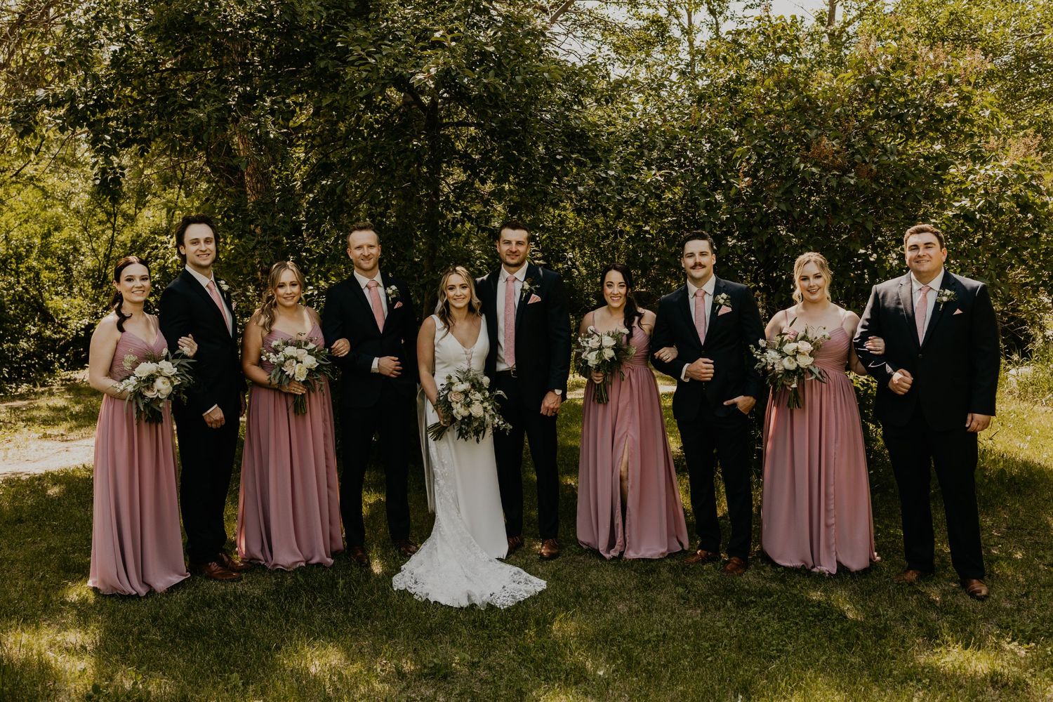 Wedding party in formal attire poses outdoors with bridesmaids in dusty rose dresses.