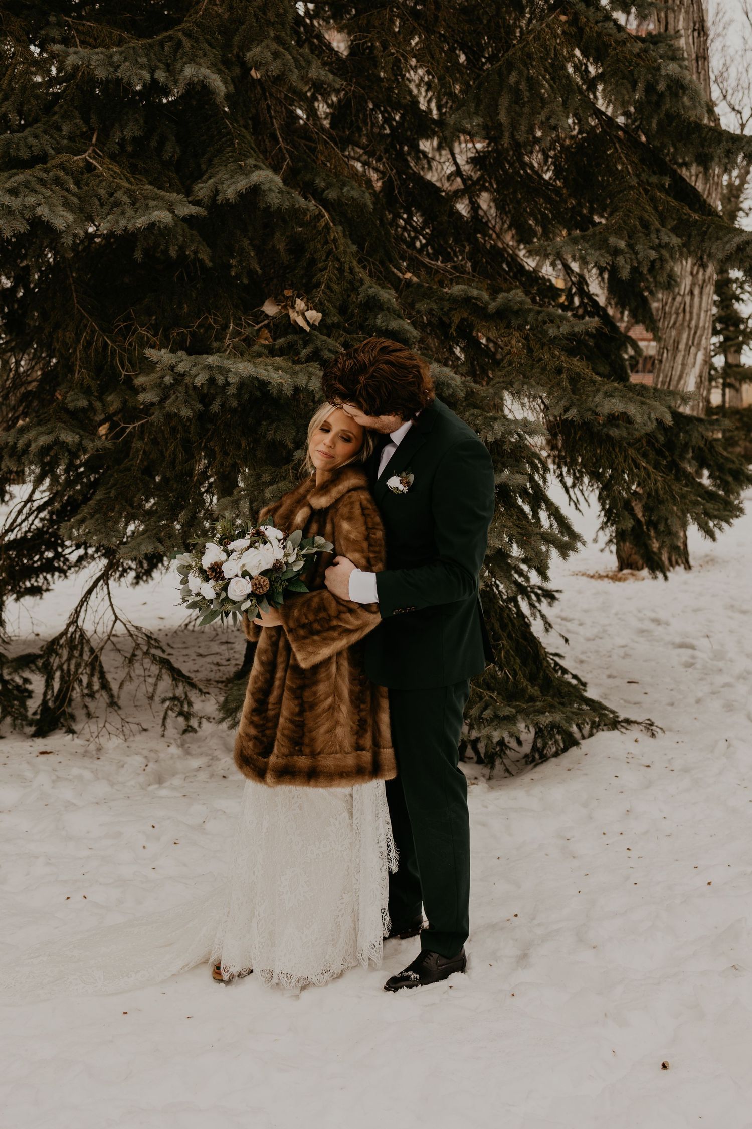 Couple shares intimate moment under pine trees in winter wedding attire.