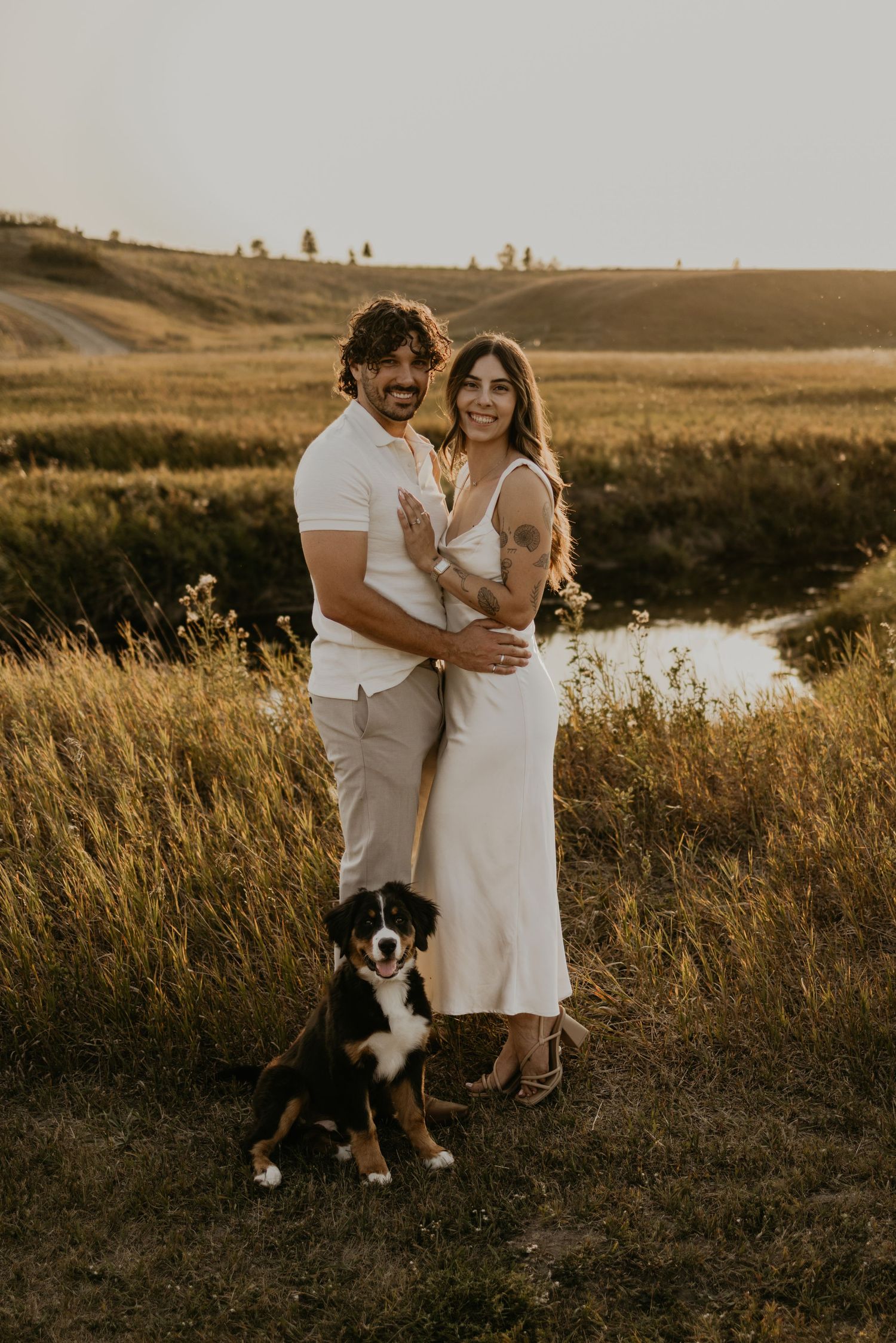 Couple in white outfits posing together in golden field at sunset with their dogs.