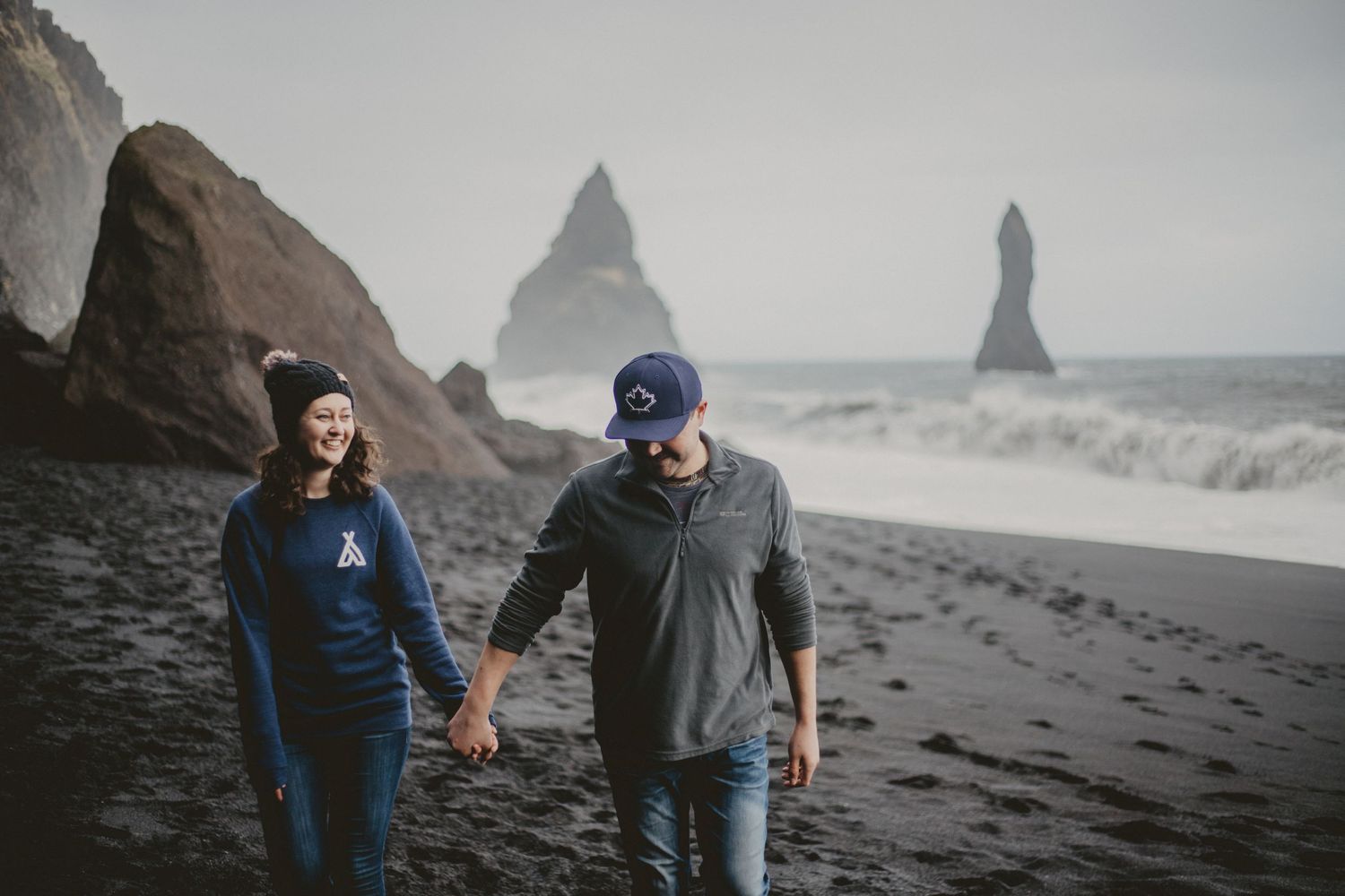 Couple holding hands while exploring a dramatic volcanic beach with ocean stacks in background.