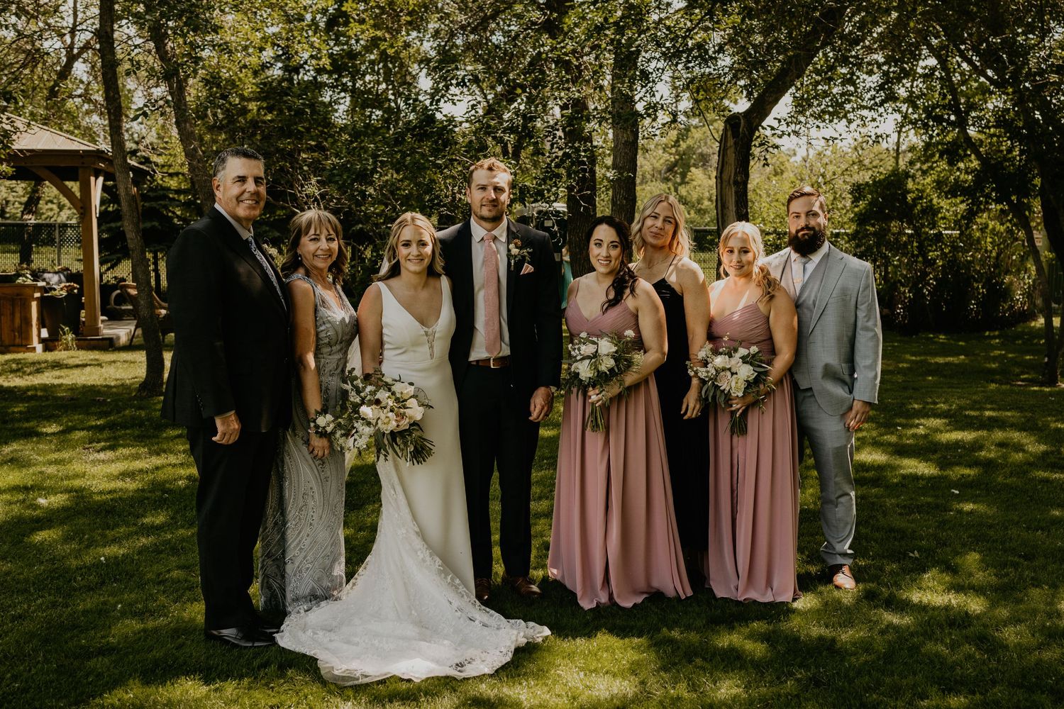 Wedding party in formal attire poses for a photo on a sunny lawn.
