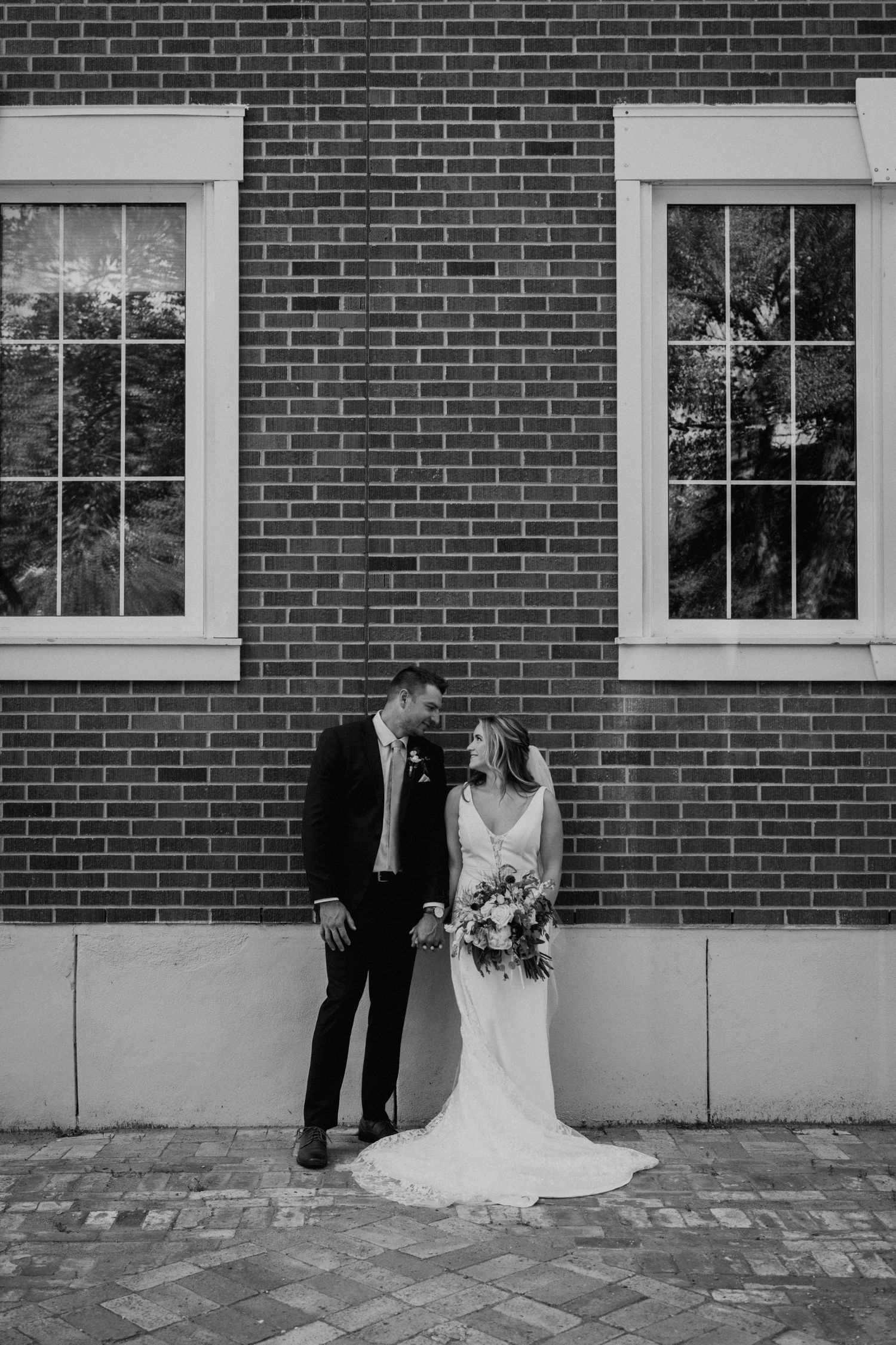 An elegant black and white portrait of newlyweds posing between two windows on a brick wall.