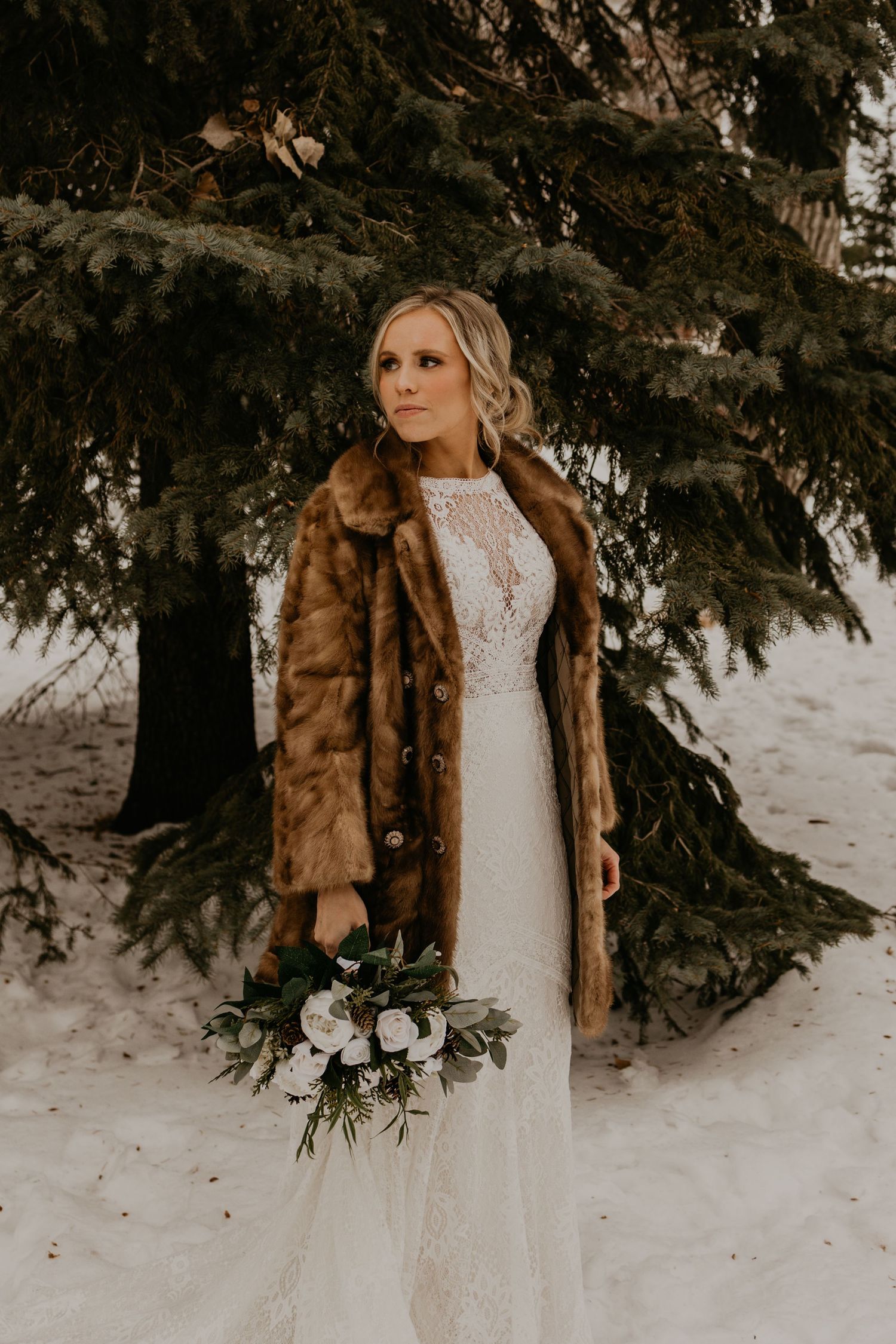 Winter bride wearing an elegant lace gown and brown fur jacket poses with bouquet against snowy evergreen backdrop.