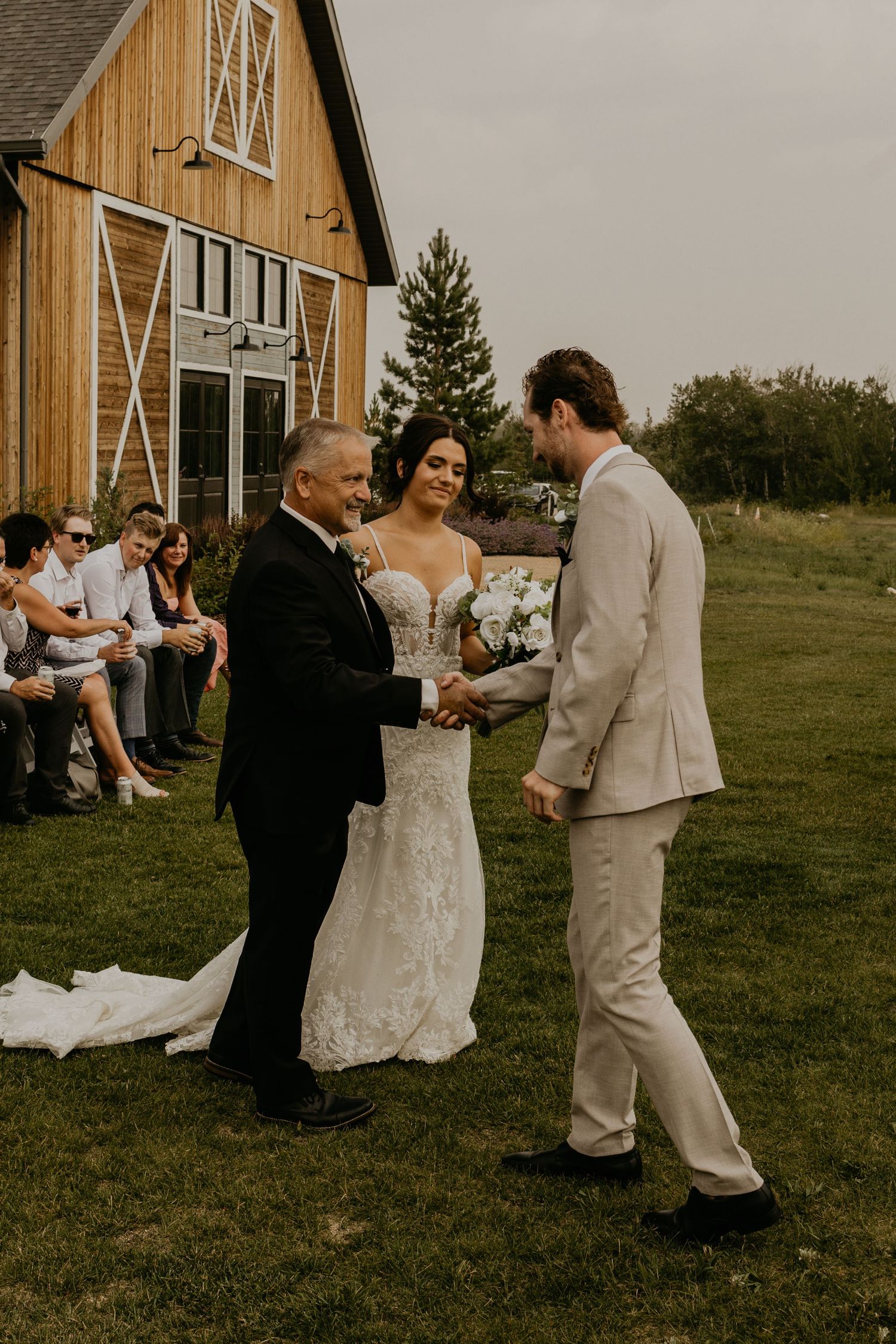 A wedding ceremony takes place in front of a rustic barn with guests seated on white chairs.