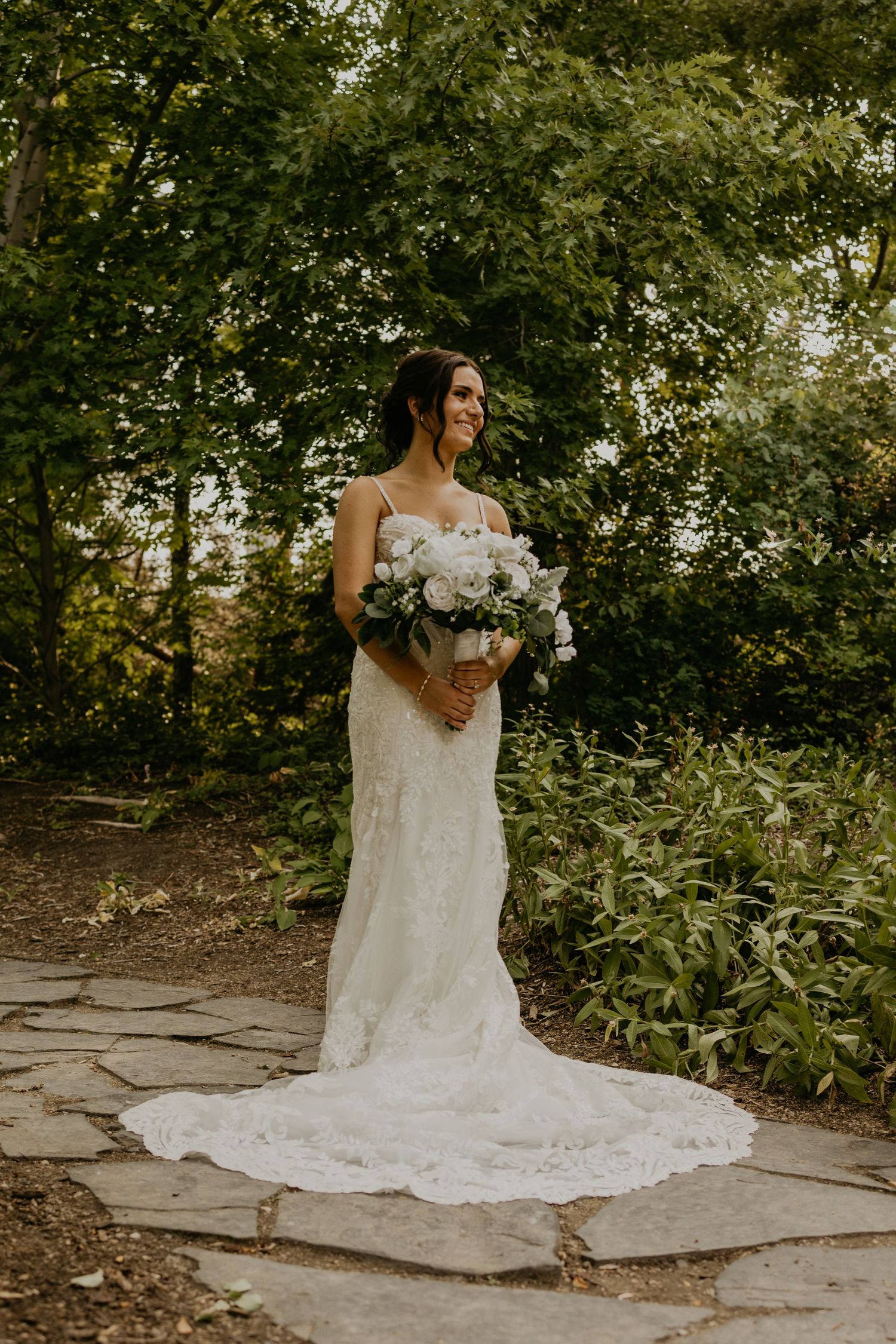 A bride in a lace wedding dress holds a white bouquet while standing on a stone path surrounded by greenery.