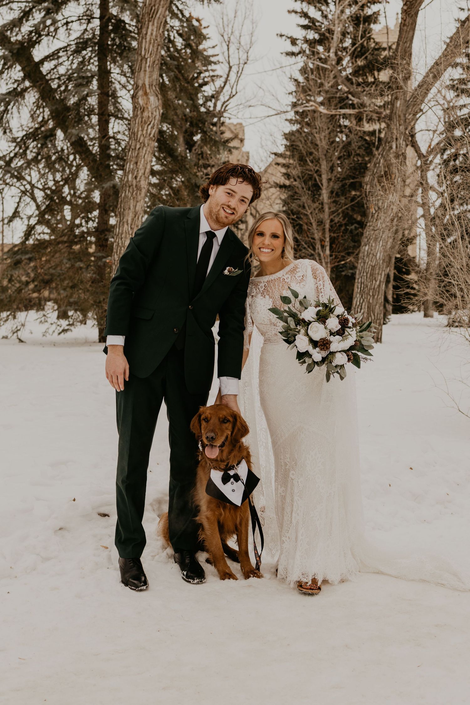 Winter wedding photo with a happy couple and their dog wearing a bow tie in snowy forest setting.