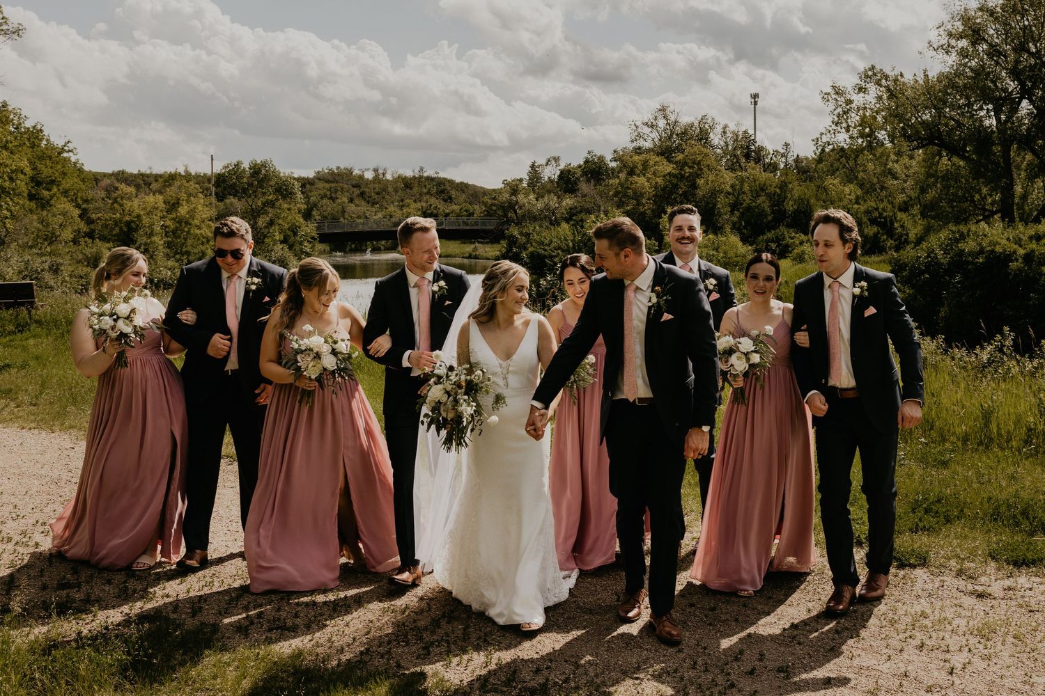 A wedding party poses on a gravel path with bridesmaids in dusty rose dresses.