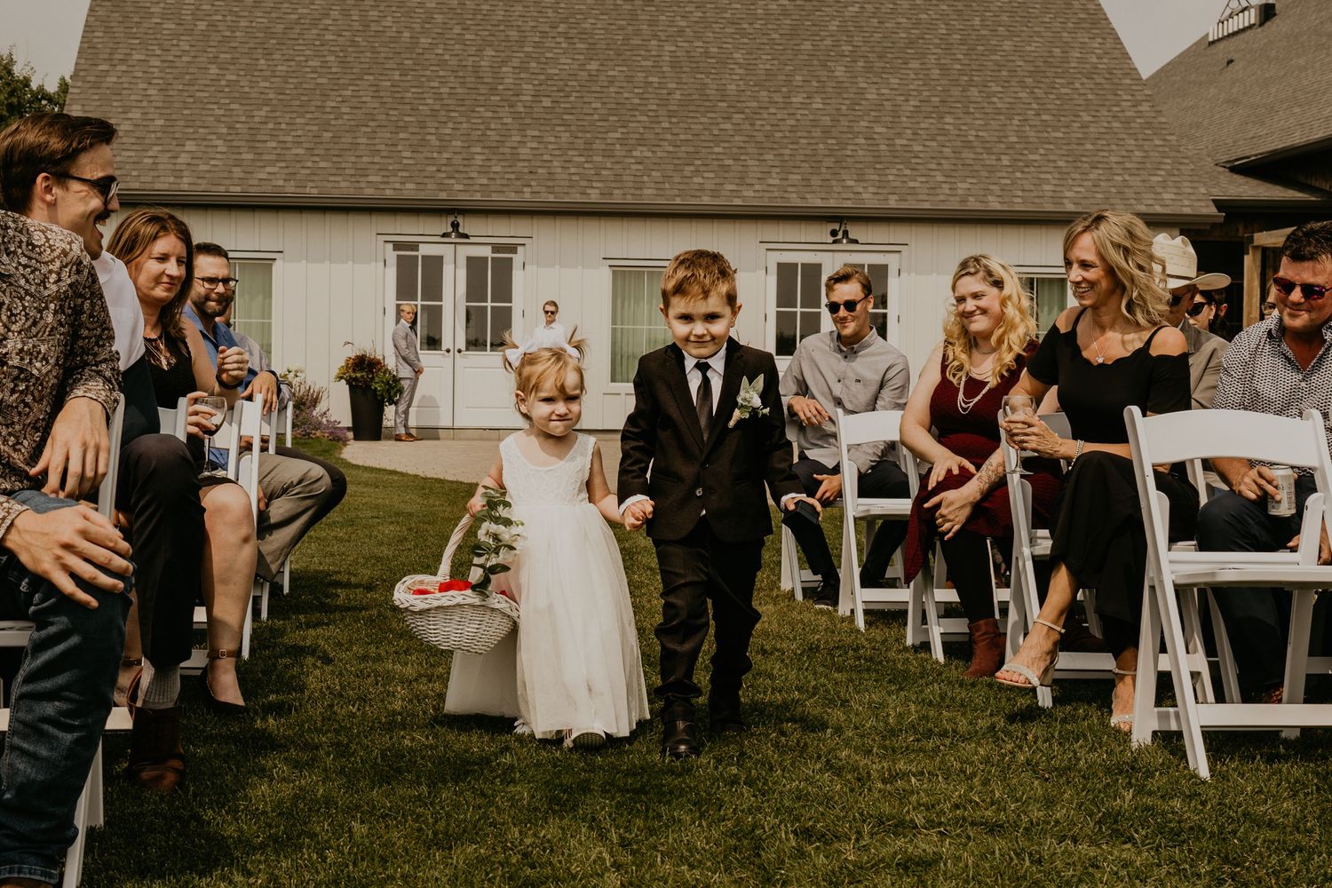 Young ring bearers walk down the aisle during a wedding ceremony with guests seated in white chairs.