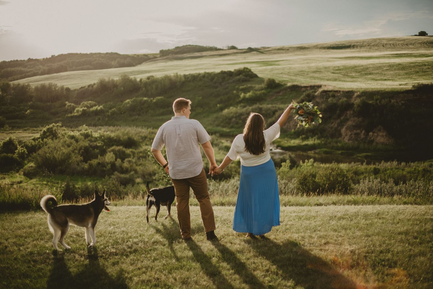 A couple and their dogs watch the sunset together in rolling green countryside.
