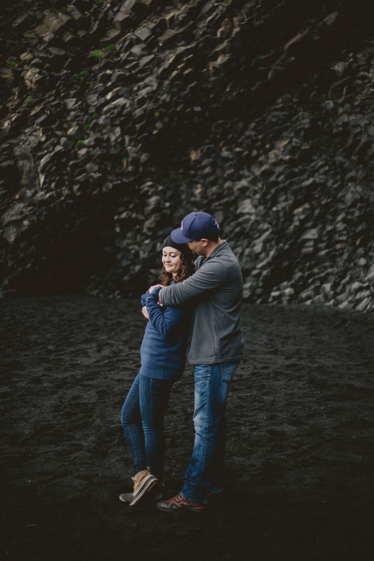 A joyful couple sharing a playful moment against a dark rocky backdrop, dressed in casual denim and grey.