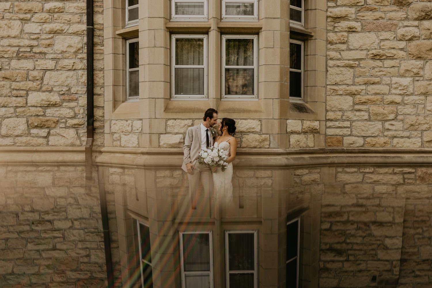 Romantic wedding portrait against stone building with window reflection.