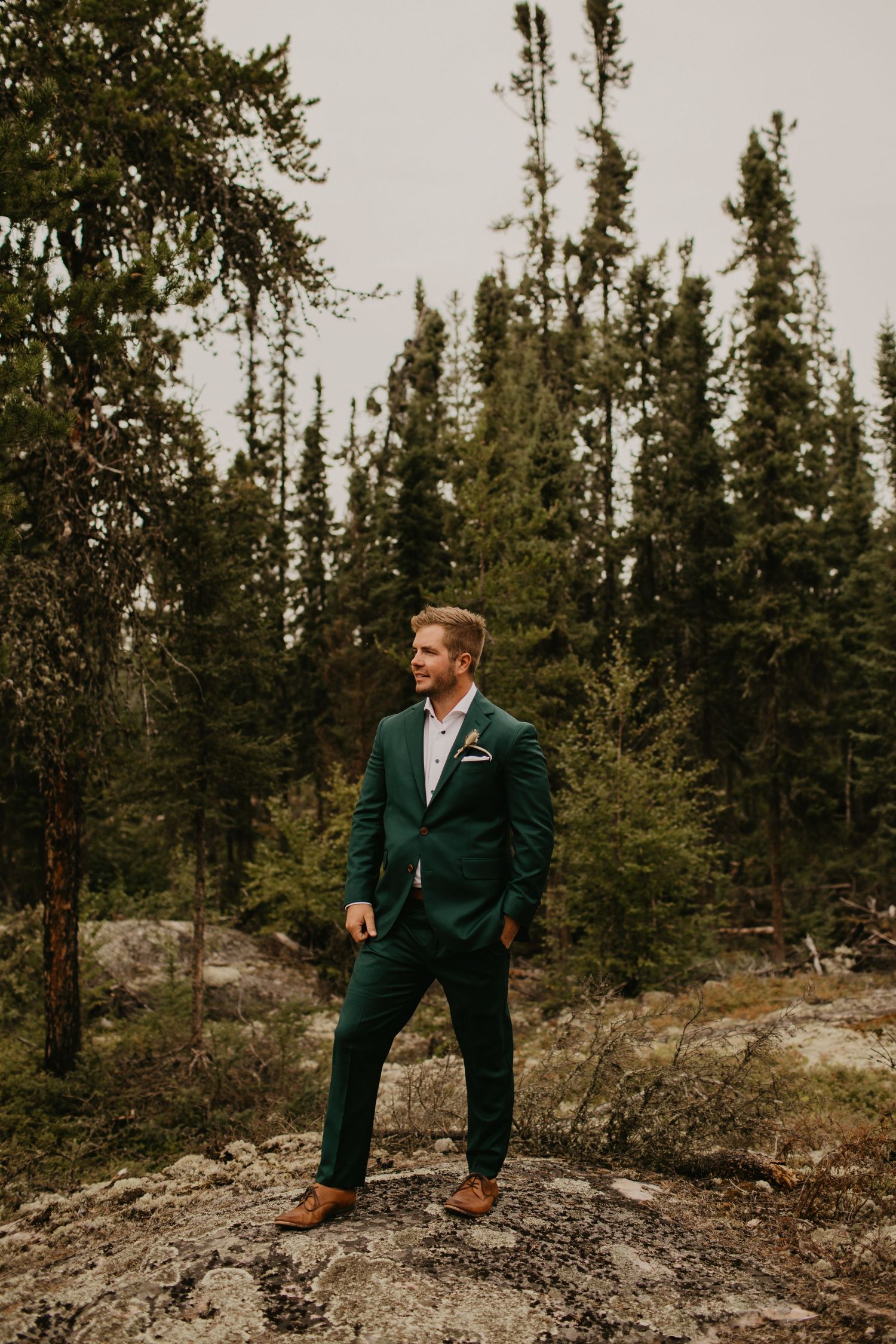 Groom in emerald green suit poses on rocky outcrop surrounded by pine forest.