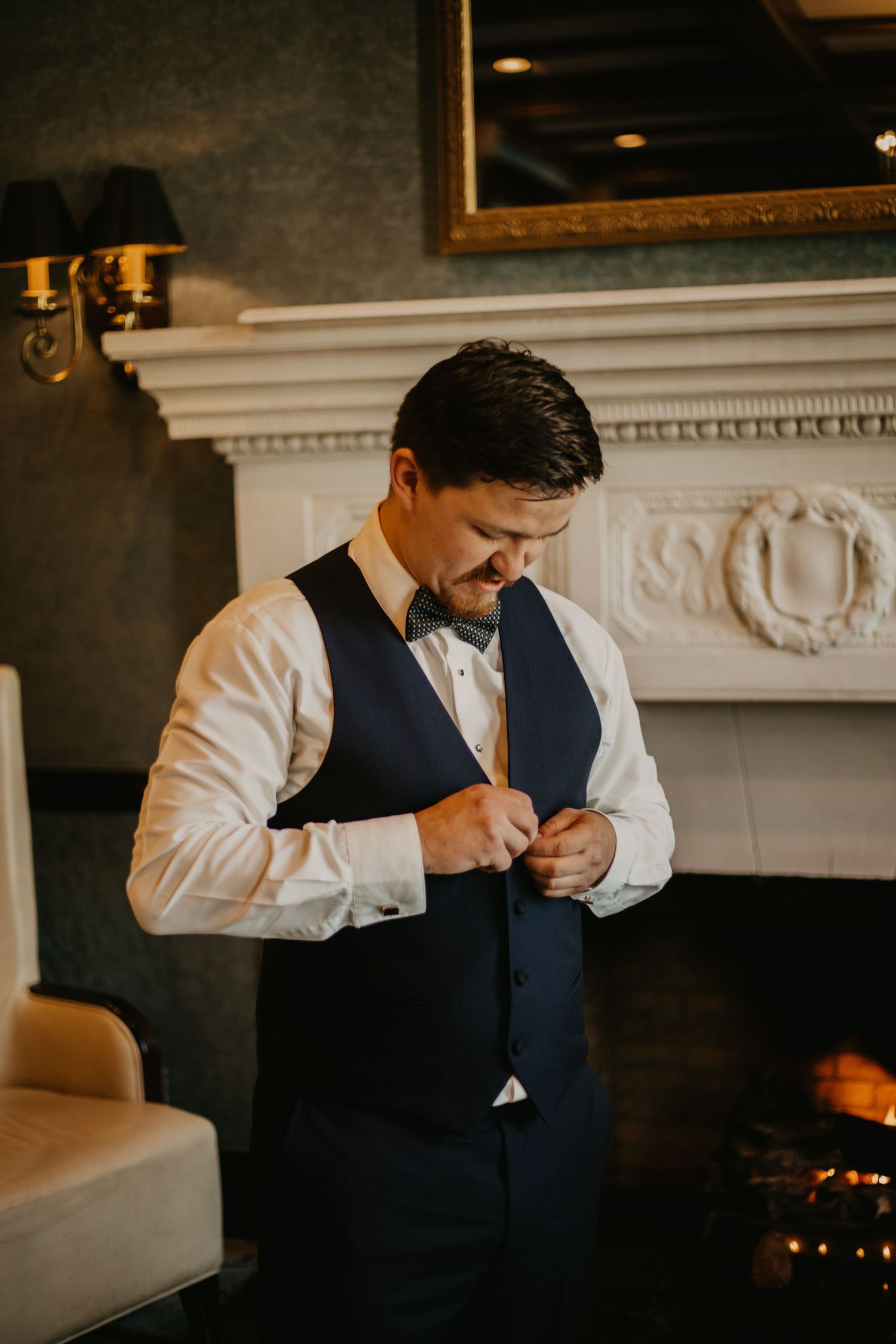 Person adjusting navy blue vest while getting dressed in formal attire near ornate fireplace.