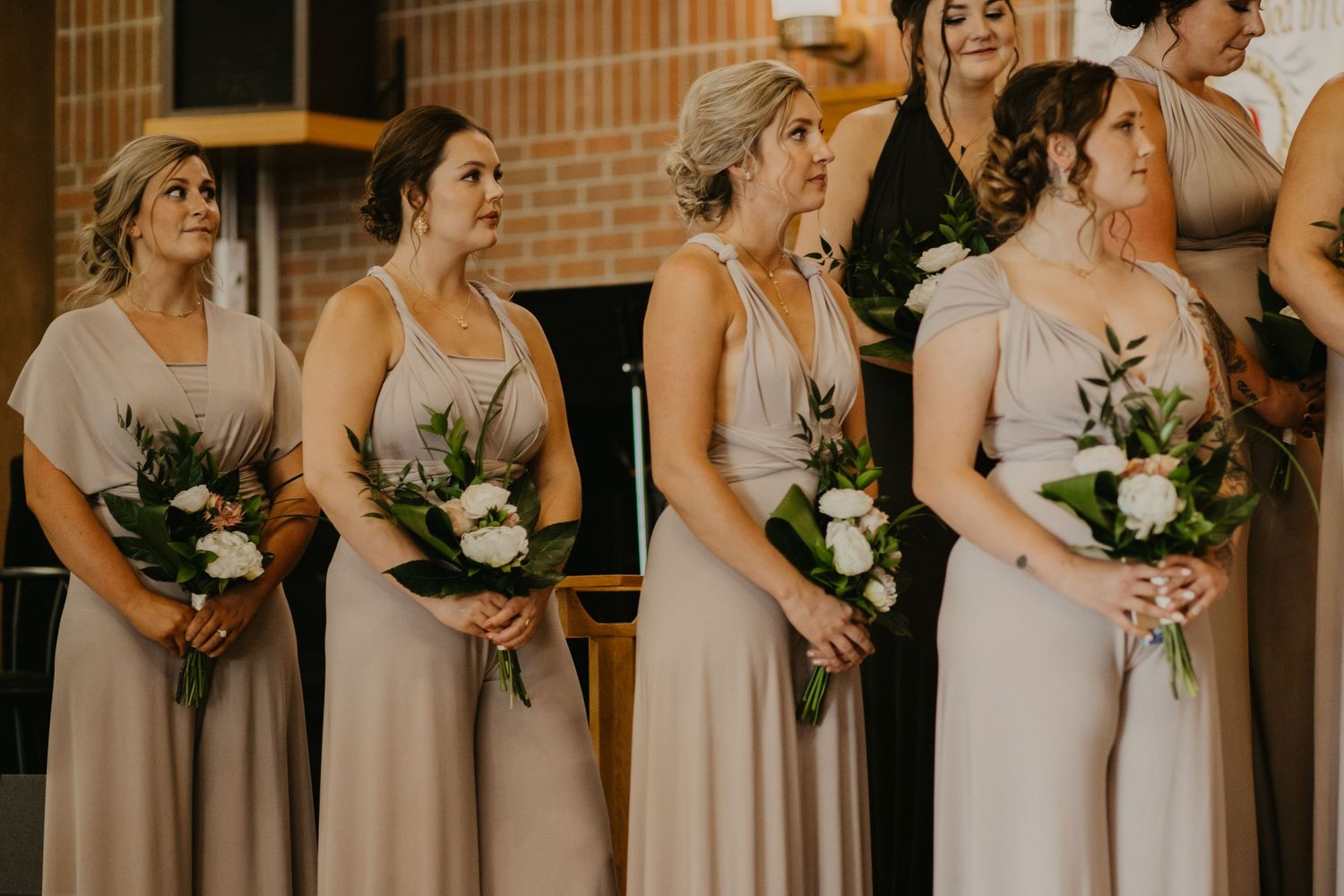 Bridesmaids in soft mauve dresses hold white bouquets while standing in a line against a brick wall.
