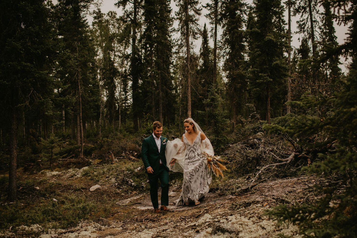Wedding couple walking together on rocky terrain surrounded by tall pine trees.