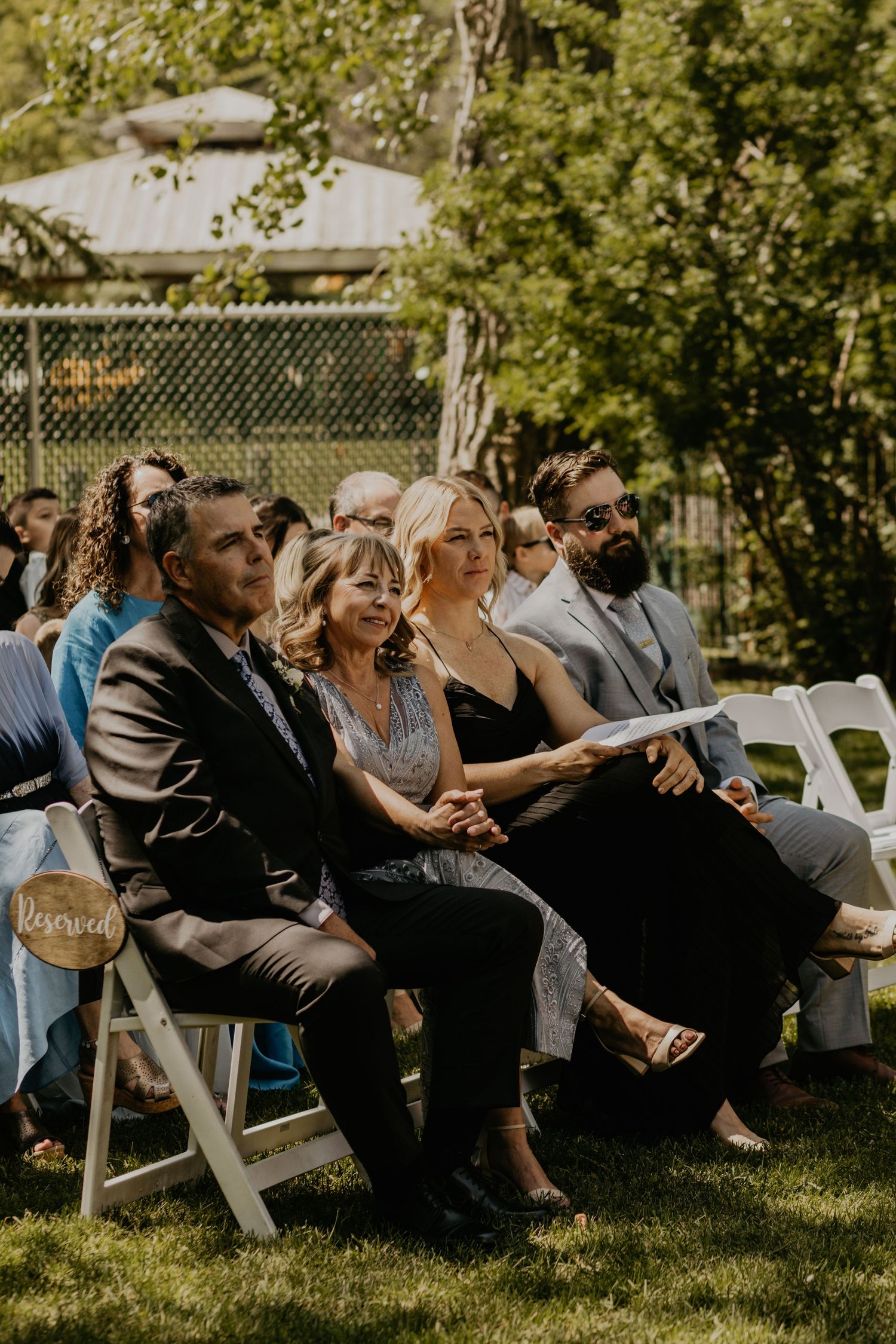 Wedding guests seated in white folding chairs on a lawn watching the outdoor ceremony.