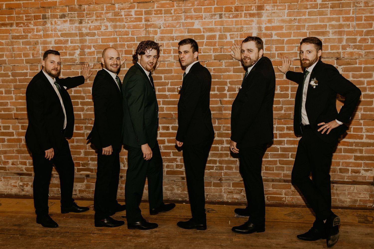 Groomsmen in black suits pose casually against rustic brick wall in wedding party photo.