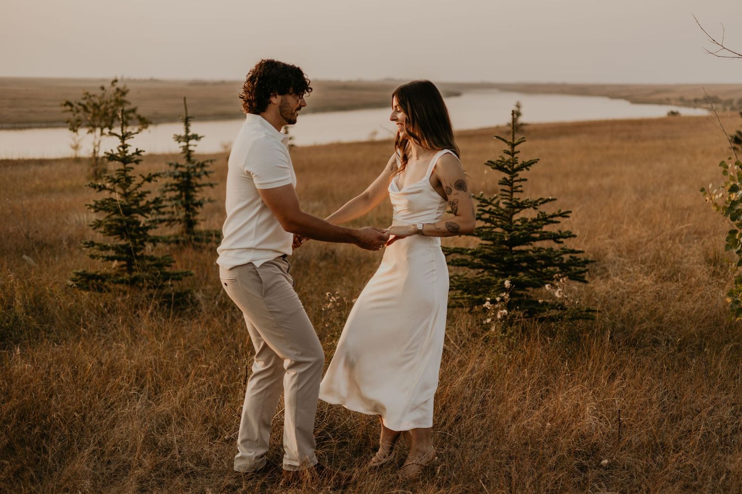 A couple embraces in a golden field at sunset with evergreen trees and a lake in the background.