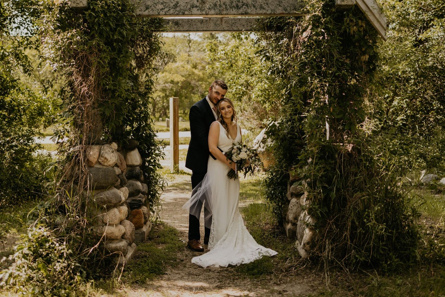 A romantic wedding couple stands beneath a vine-covered stone archway in a garden setting.